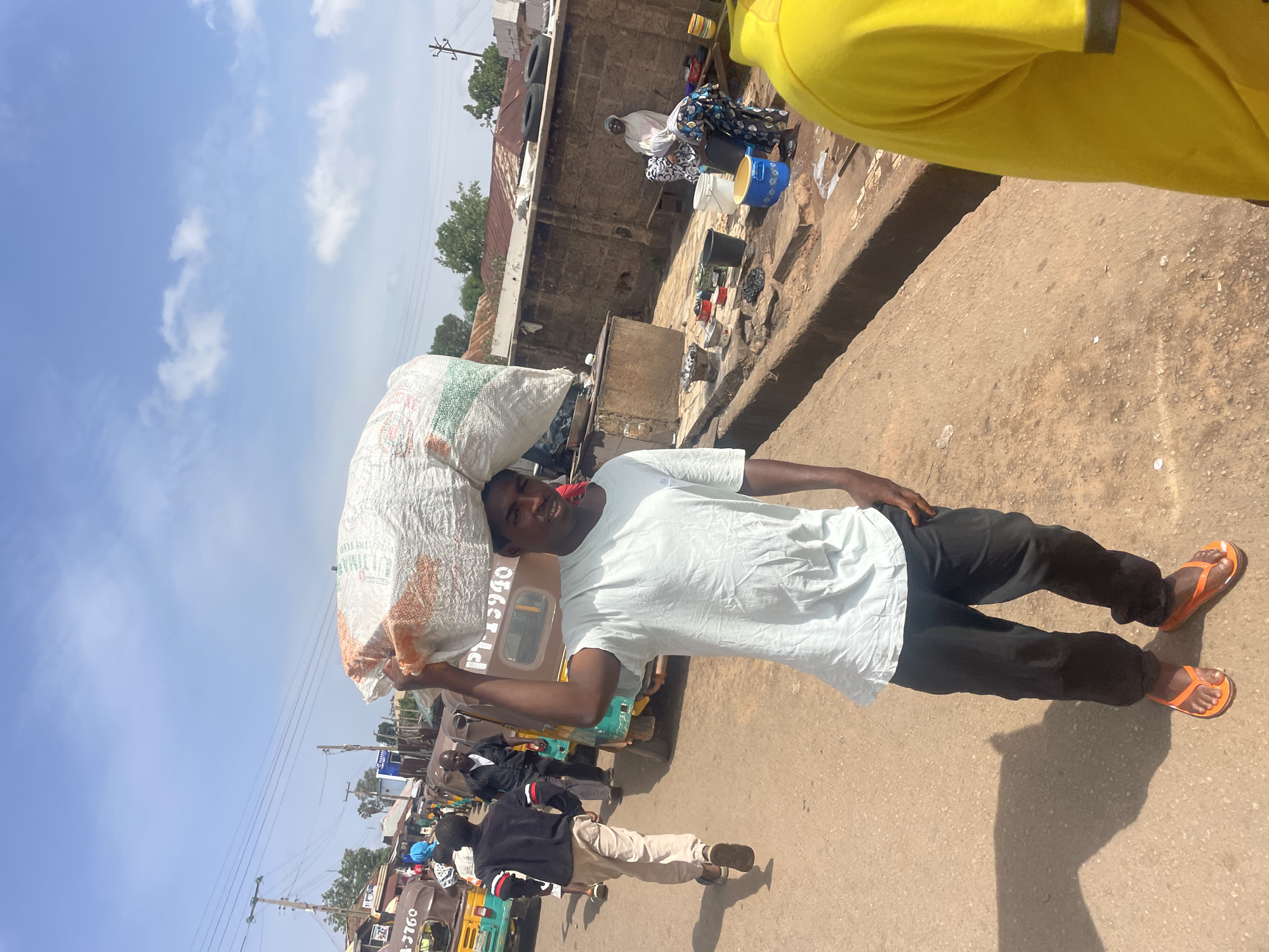 A man carries a large white sack on his head in Gura Top, Nigeria. He wears a light blue t-shirt, dark pants, and orange sandals. The street scene includes other pedestrians, auto rickshaws, and buildings. Some people are washing clothes. The sky is partly cloudy.