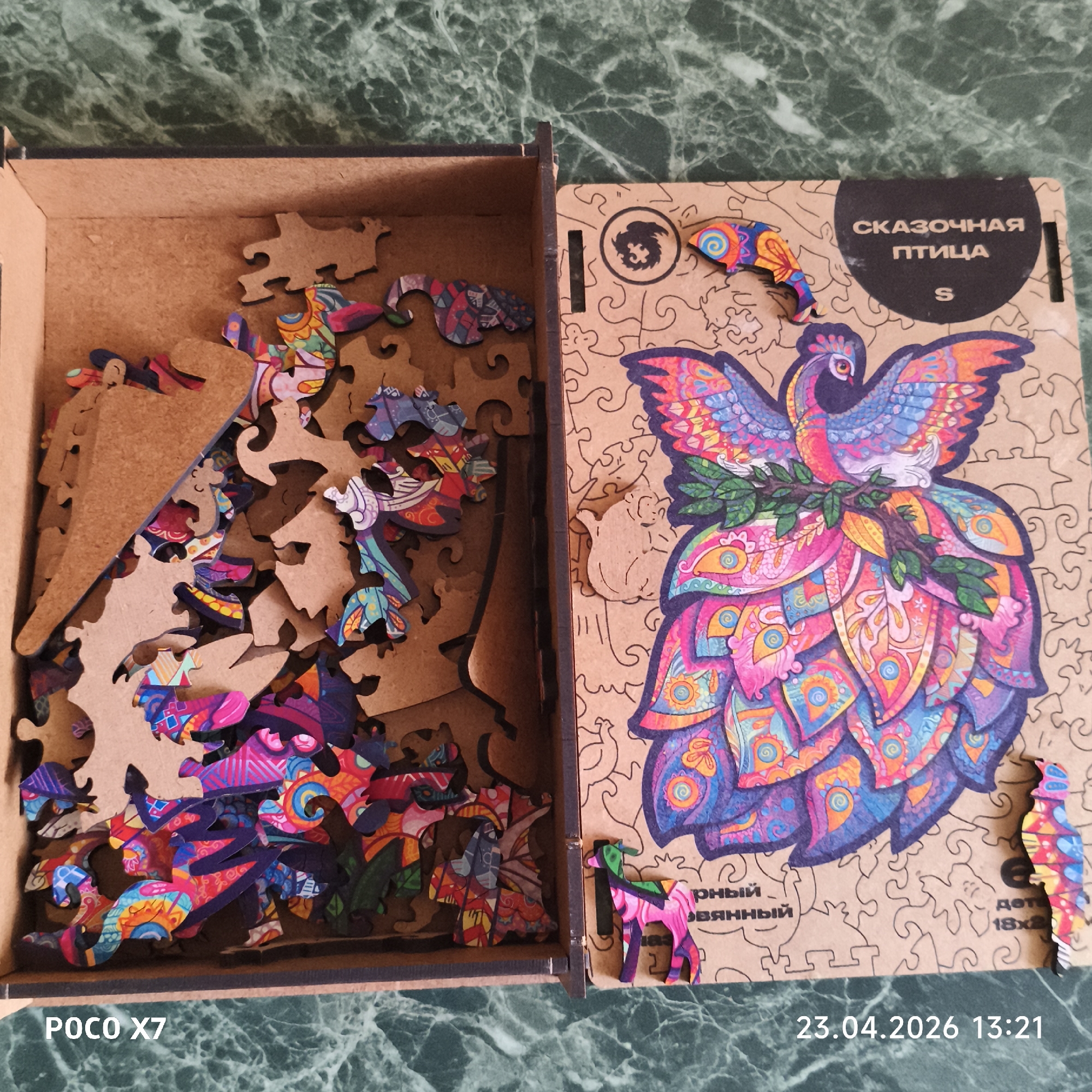 An overhead shot displays an open wooden puzzle box and its lid resting on a dark green, marble-patterned surface, possibly a countertop. The scene suggests the puzzle is either new, being prepared for assembly, or has recently been disassembled.
The puzzle's central image, depicted on the lid and on the colored pieces, is a vibrantly stylized bird, possibly a mythical creature or a peacock. Its body and wings are adorned with intricate patterns in rich shades of pink, blue, orange, and purple, giving it a whimsical and colorful appearance. The bird is shown holding a small branch with green leaves.
The puzzle pieces themselves are crafted from light-brown wood, visible on their unprinted reverse sides. They are distinctive for their irregular, non-traditional shapes, diverging from standard jigsaw cuts. Many pieces are cut into recognizable figures, such as a small horse-like animal, a fish or bird-like piece, and other unique animal or abstract forms, adding to the puzzle's unique character.
Inside the open wooden box, a collection of puzzle pieces is visible. Some pieces remain partially connected to the wooden frame they were originally cut from, while others are loose and scattered within the box. The box lid, lying flat beside the open box, functions as a visual guide, presenting the completed bird image alongside outlines of some individual pieces on its light-brown wooden surface.
Visible text on a black circular label on the lid reads in Cyrillic: "СКАЗОЧНАЯ ПТИЦА" (Fairy Bird), followed by "S", likely indicating the puzzle size. Partially visible Cyrillic text below the main image reads "ОРИГИНАЛЬНЫЙ ДЕРЕВЯННЫЙ" (Original Wooden), and "дет 18x2", possibly referring to age or dimensions. Watermarks in the bottom corners of the image display "POCO X7" (the device used to capture the image) and "23.04.2026 13:21" (date and time). The image is located in Chaikovsky, Russia.