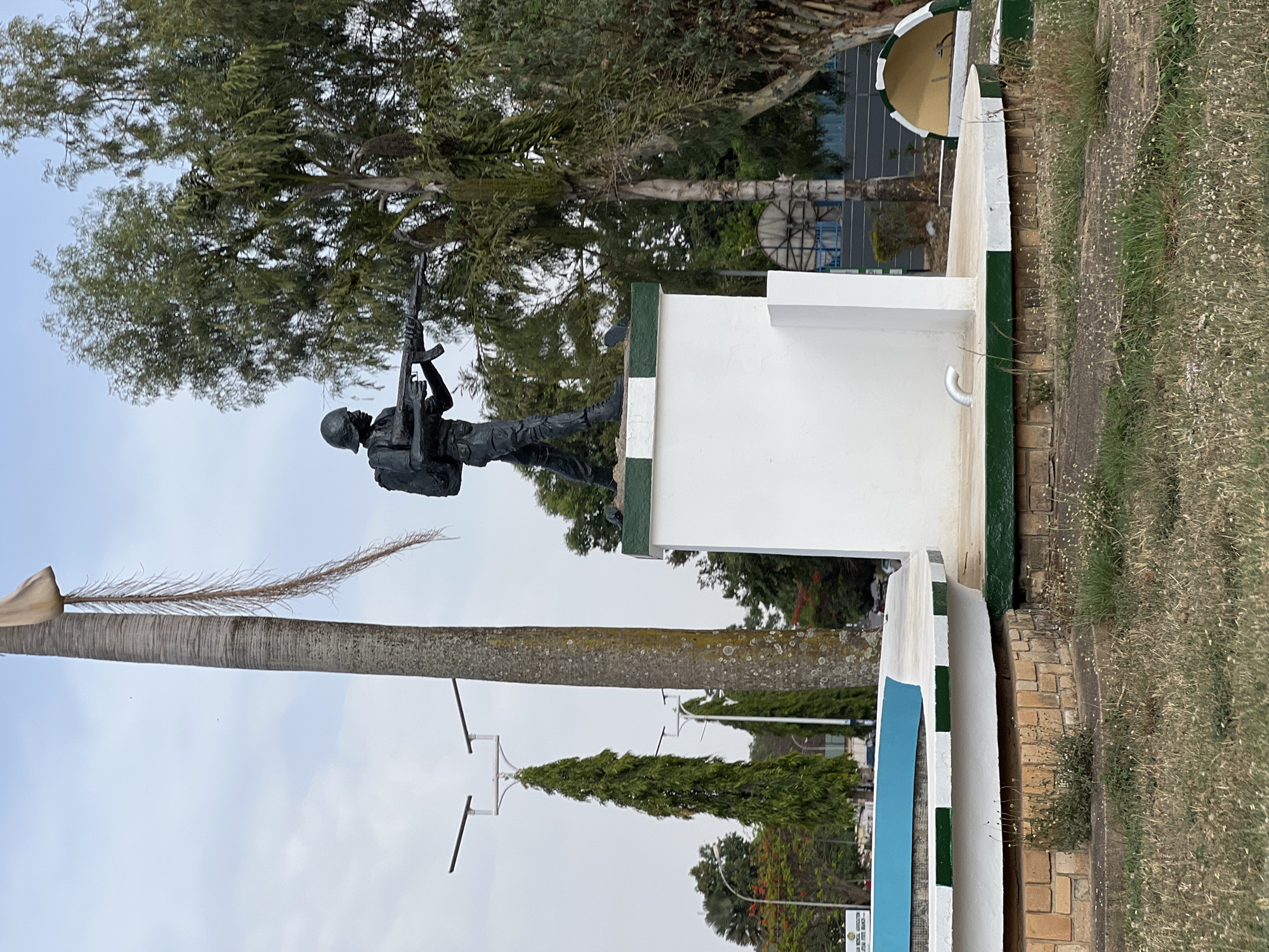 The image shows a statue of a soldier with a rifle, located on a white painted pedestal with green trim. The statue is in front of a tree. The statue is on the grounds of the Maxwell Khobe Cantonment in Jos, Nigeria. The surrounding environment includes trees, grass, and a building.