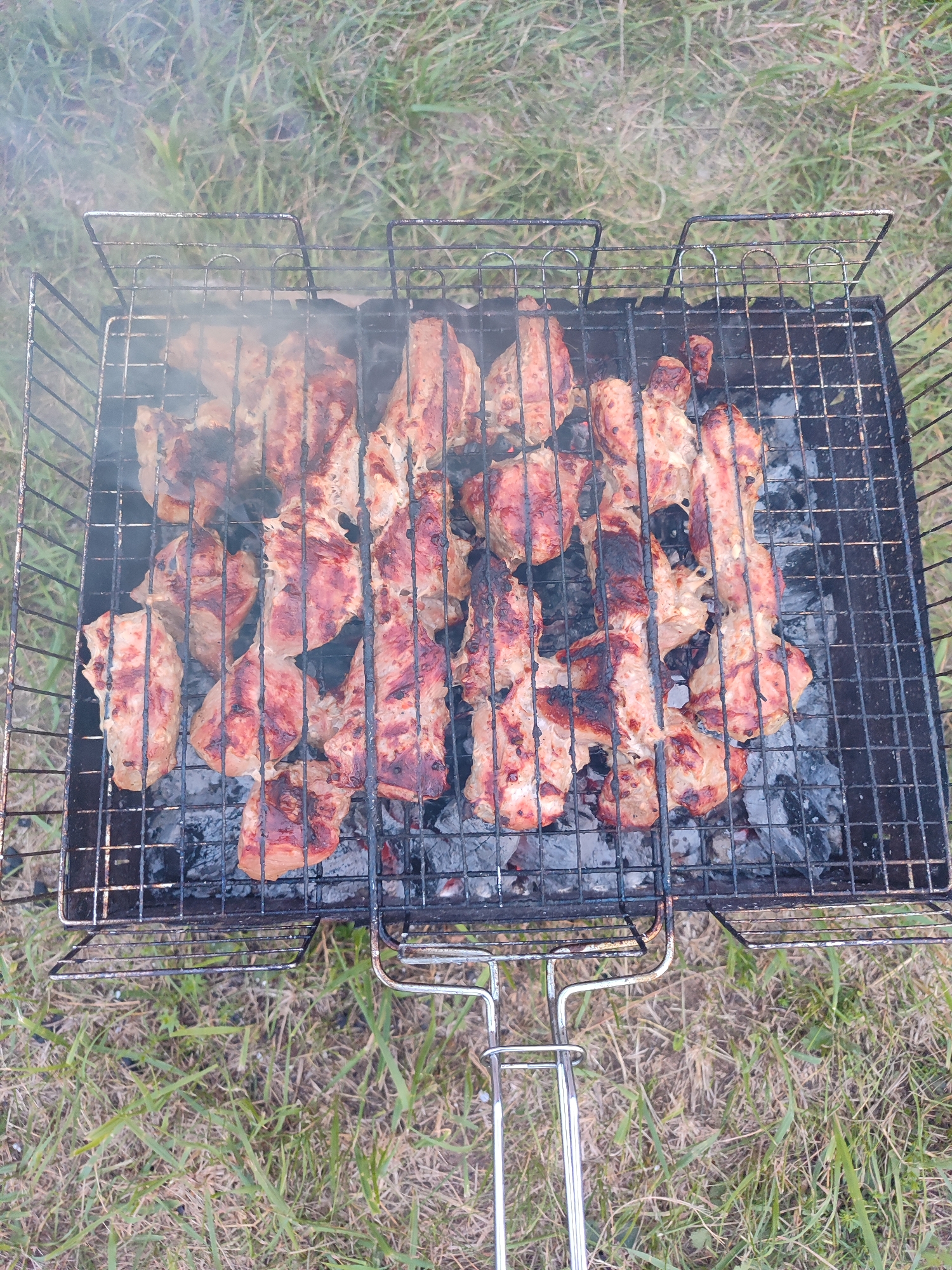The image shows a close-up, overhead view of a metal barbecue grill filled with pieces of meat being cooked over hot coals. The meat appears to be pork shashlik, with some pieces showing charring from the heat. Smoke is rising from the grill. The grill is situated on a patch of dry grass, suggesting an outdoor setting. The image does not contain any elements that would indicate its specific location within the city of Chaikovsky, Russia. There are no people or any identifying landmarks visible.