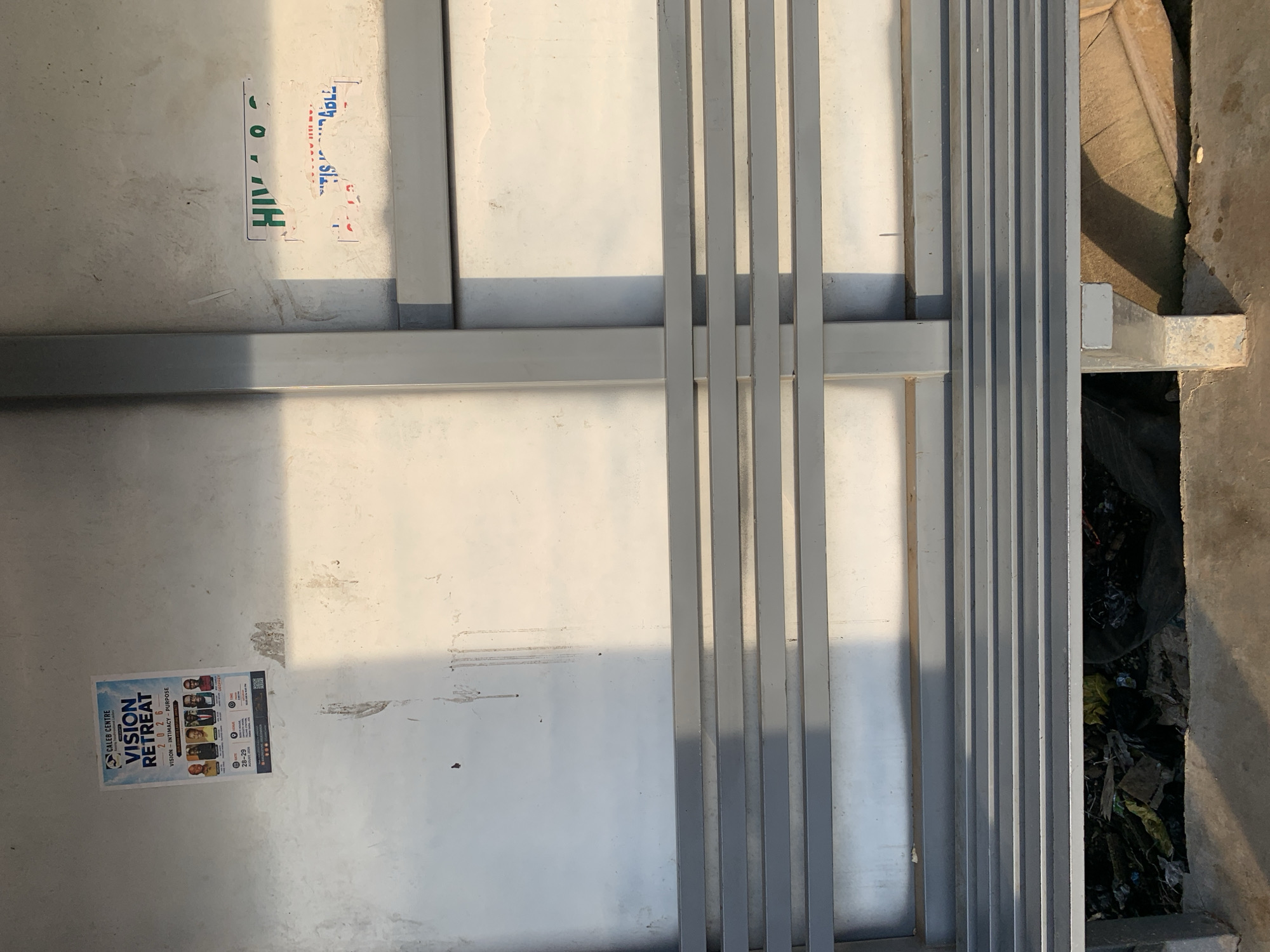 The image depicts an outdoor scene, likely a bus stop or waiting area, in Rafin Par, Nigeria. A metal bench with multiple horizontal slats runs across the lower half of the frame. Behind the bench, a light-colored, textured wall serves as a backdrop. On the wall, two posters are visible. The left poster, featuring a light blue and white design, advertises a "CALEB CENTRE VISION RETREAT 2026" with the tagline "VISION - INTIMACY - PURPOSE". It displays portraits of five individuals. To the right of this poster, another poster or sign with green and white lettering is partially visible, with the words "HIV O C" and a portion of another phrase below it. The area beneath the bench is cluttered with debris, including plastic bags and other refuse, suggesting an unkempt environment. Sunlight casts shadows across the scene, indicating it is daytime.