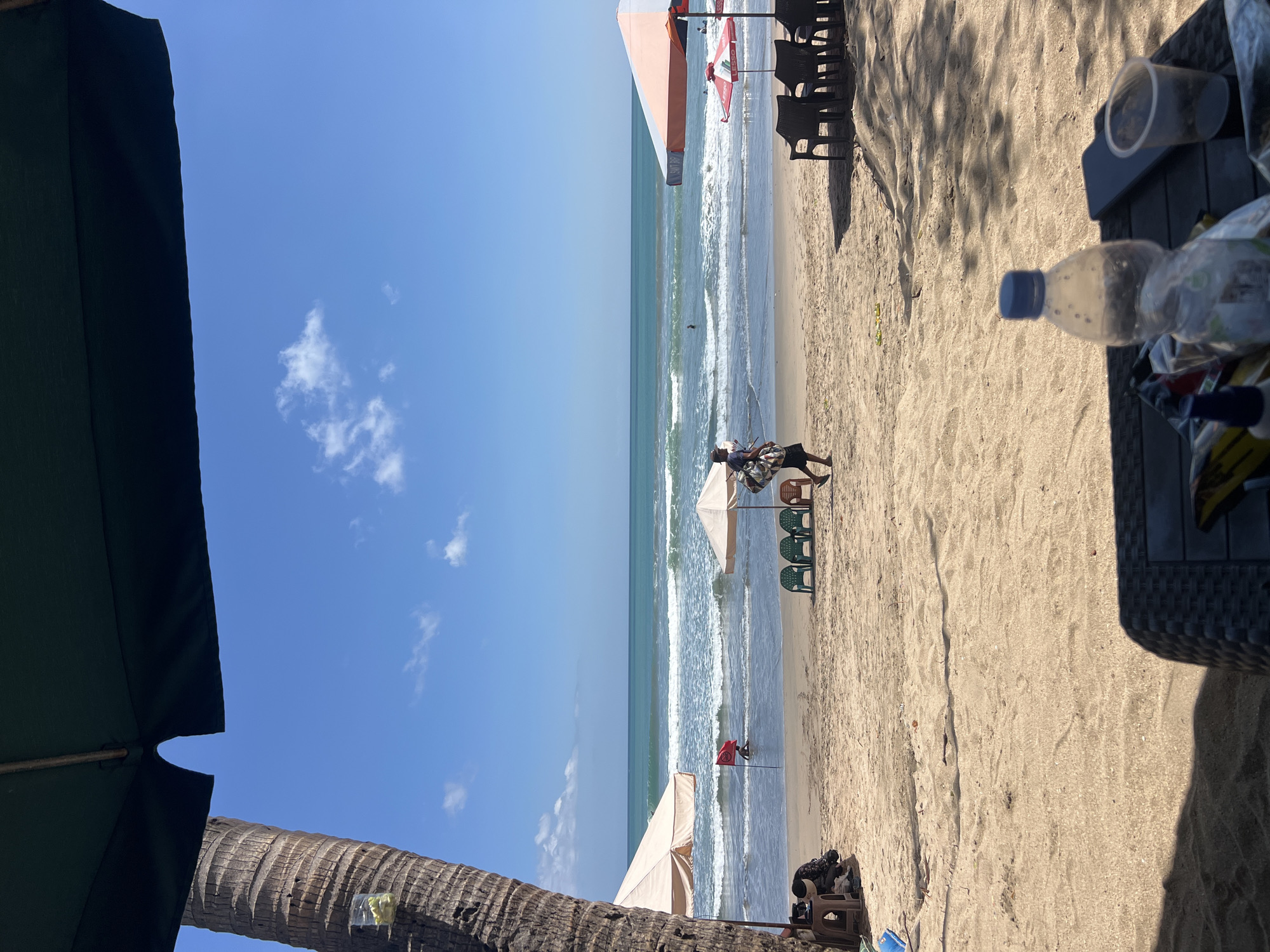 The image depicts a beach scene in Kuta, Indonesia. In the foreground, a table with a plastic bottle, a smartphone, and a cup is visible, casting shadows from a dark umbrella overhead. The beach itself is sandy, with a few green chairs and a person walking to the right. Further back, several beach umbrellas are set up, with people relaxing under them. The ocean is visible in the background, with gentle waves breaking on the shore and a few people swimming or wading in the water. A red flag is planted in the water, indicating a potential warning. A tall palm tree stands to the left of the frame, adding to the tropical ambiance. The sky is a clear blue with a few white clouds.
