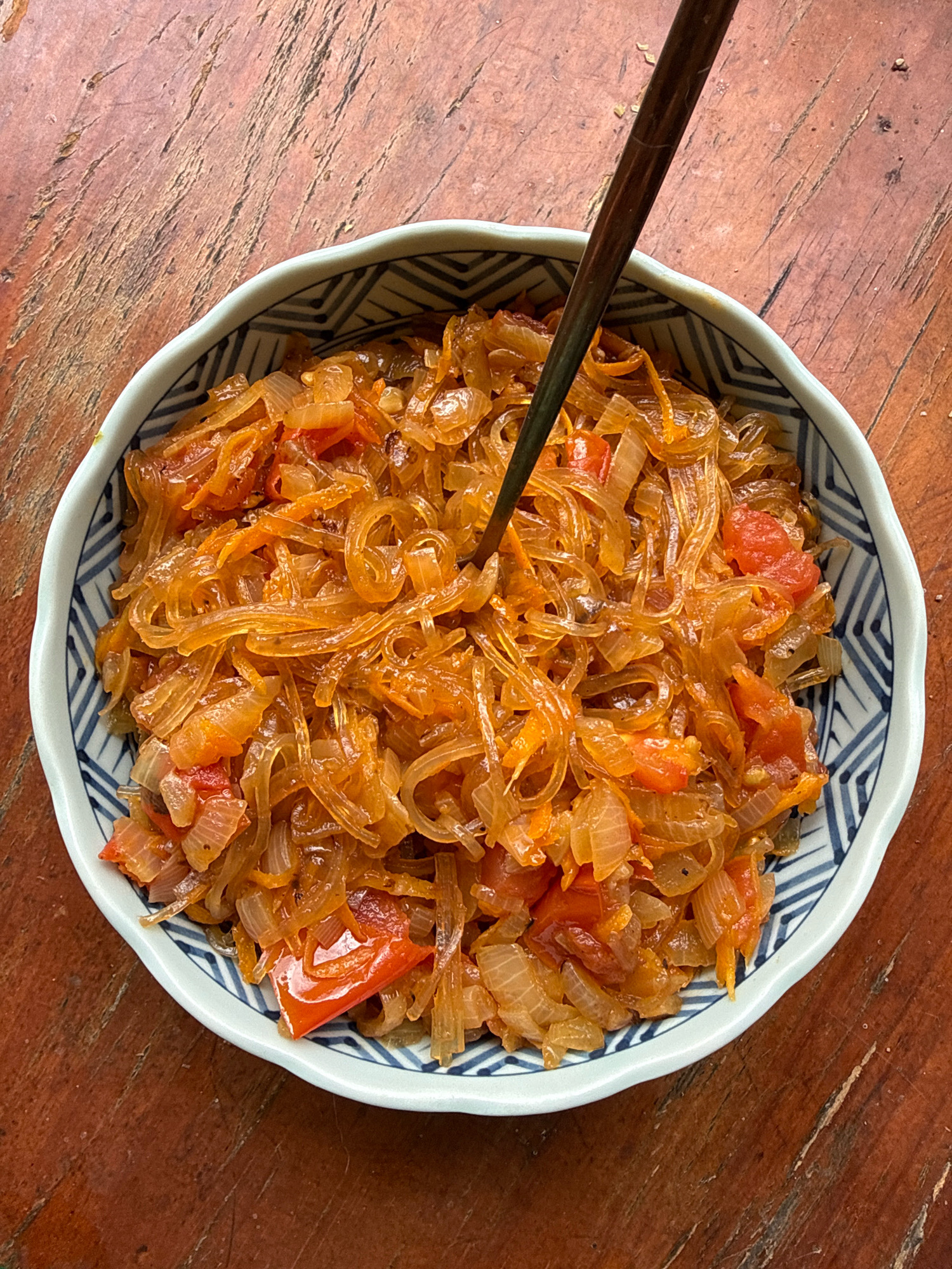 A close-up, overhead shot shows a bowl of stir-fried glass noodles with onions and tomatoes, positioned on a wooden table. A dark wooden chopstick is inserted into the food, suggesting it is being eaten. The bowl has a white and blue geometric pattern around the rim. The wooden table has a visible grain and appears to be outdoors, possibly in Hiệp Thạnh, Vietnam, given the warm lighting. There are no people visible in the image.