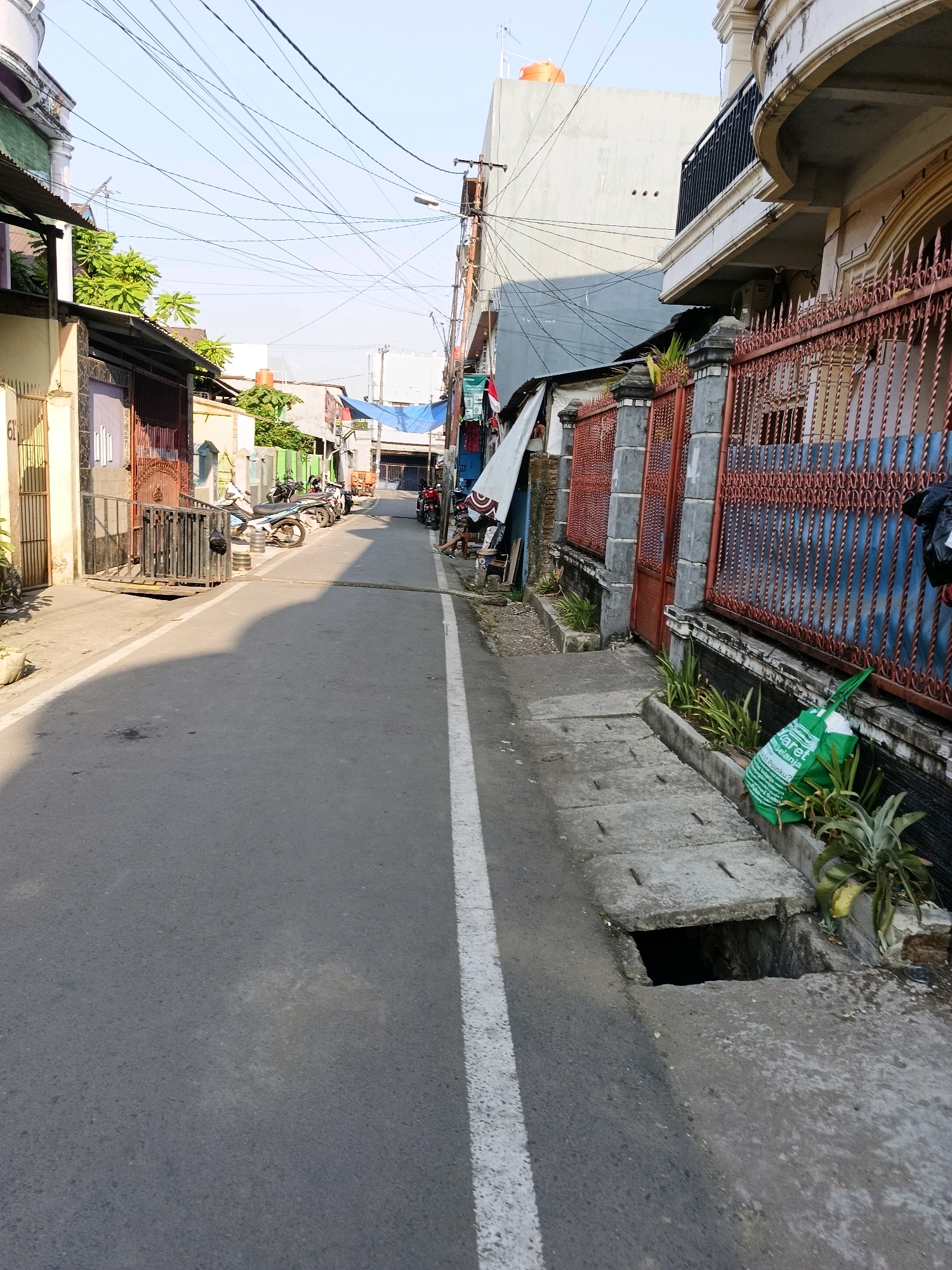 The image depicts a street scene in Makassar, Indonesia. The road is paved and has a white dividing line. On the left side of the road, there are several parked motorcycles, some with awnings providing shade. Buildings with various architectural styles line both sides of the street, some with ornate fences and balconies. A storm drain is visible on the right side of the road, with an open grate. A green plastic bag with writing on it is placed near the storm drain. Overhead, a complex network of electrical wires crisscrosses the sky. The overall environment appears to be an urban residential or commercial area, with a moderate amount of clutter and signs of daily life. The lighting suggests it is daytime, with the sun casting shadows on the road.