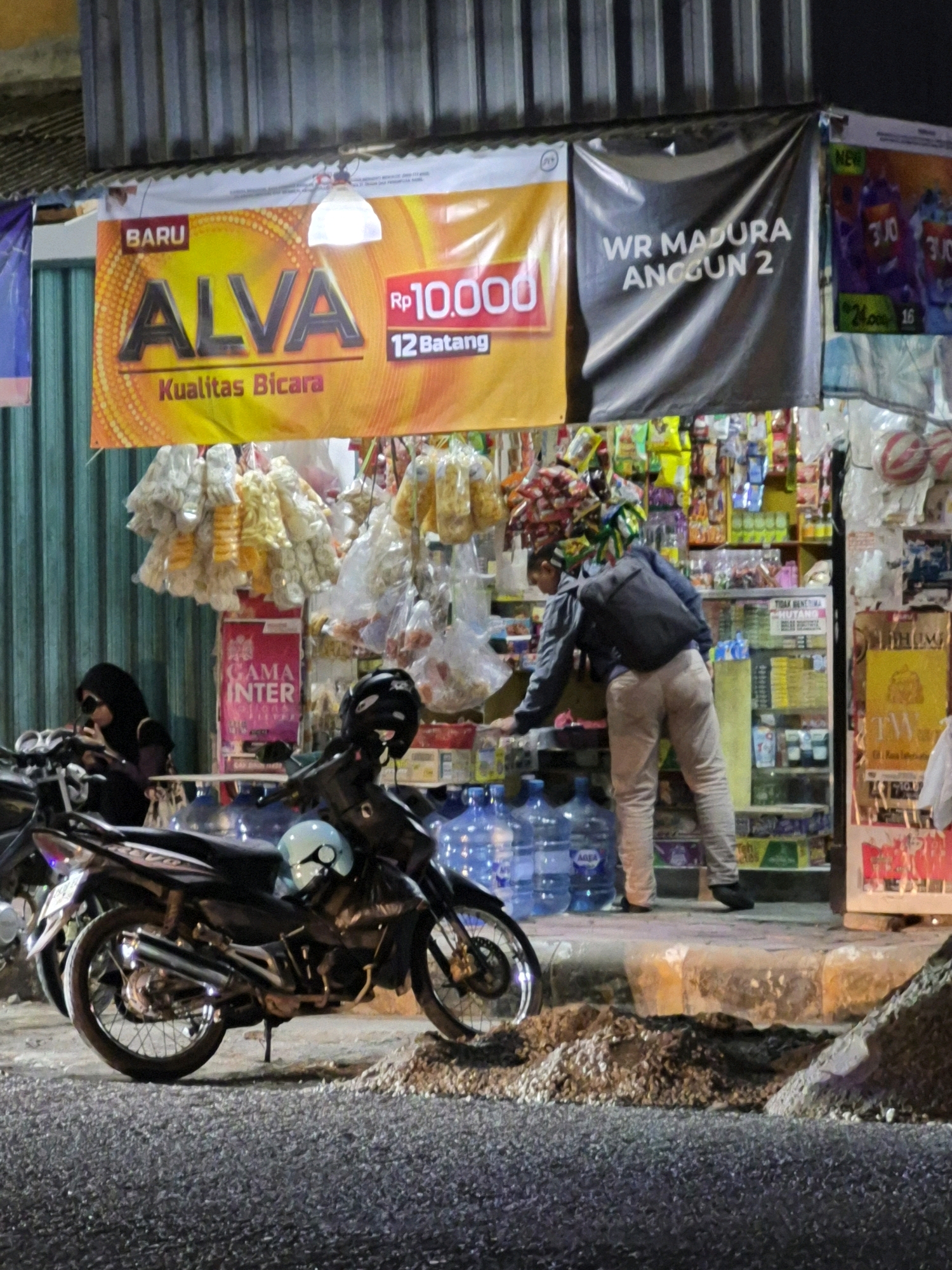 The image captures a street-level view of a roadside store in Purworejo, Indonesia, at night. In the foreground, two motorcycles are parked on a gravel surface. A woman wearing a black hijab and sunglasses is seated on the left motorcycle, looking at her phone. A blue helmet rests on the seat of the central motorcycle.
Behind the motorcycles, the store's facade is visible, featuring several large banners. The leftmost banner is yellow and orange with the text "BARU ALVA Kualitas Bicara" and a price of "Rp 10,000 12 Batang". To its right is a black banner with white text that reads "WR MADURA ANGGUN 2". Further to the right, parts of other banners with product advertisements are visible.
The store itself appears to be a small convenience store or "warung", with shelves stocked with various goods, including bottled water and packaged snacks, visible through a glass display case. A man wearing a grey jacket and light-colored pants is seen bending over, reaching into the display case. Large blue water jugs are stacked outside the store, near the entrance. The ground in front of the store is a mixture of gravel and what appears to be a paved sidewalk or curb. The lighting suggests it is nighttime, with artificial lights illuminating the scene.