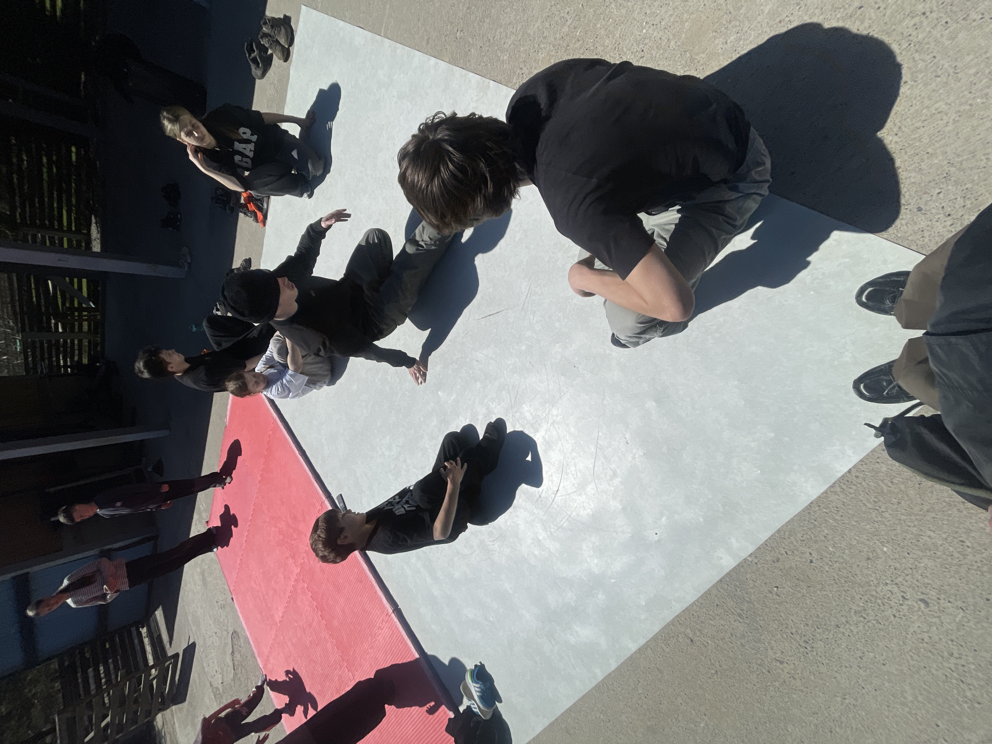 A medium-angle shot reveals a group of approximately ten people gathered on a large rectangular surface that is red on one side and gray on the other. The individuals are arranged in a semi-circle, with some kneeling or crouched down and others standing. Most are wearing dark clothing. The group is assembled outdoors, likely in the city of Daurskoe, Russia. The light source seems to be coming from overhead, casting shadows behind the people.