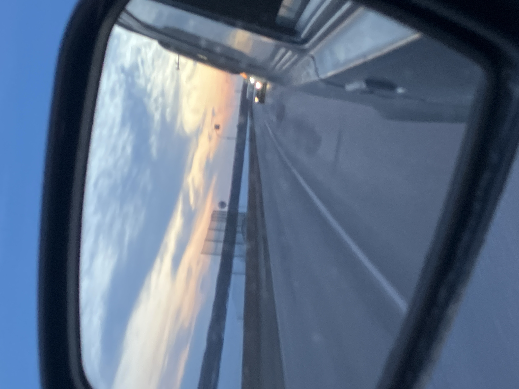 The image shows a side view mirror reflecting the road and the sky during sunset. The road is a multi-lane highway. In the distance, there are car lights. The sky is a mix of blue, white, and orange colors. The location cannot be determined as Noviy, Russia, with the given information.