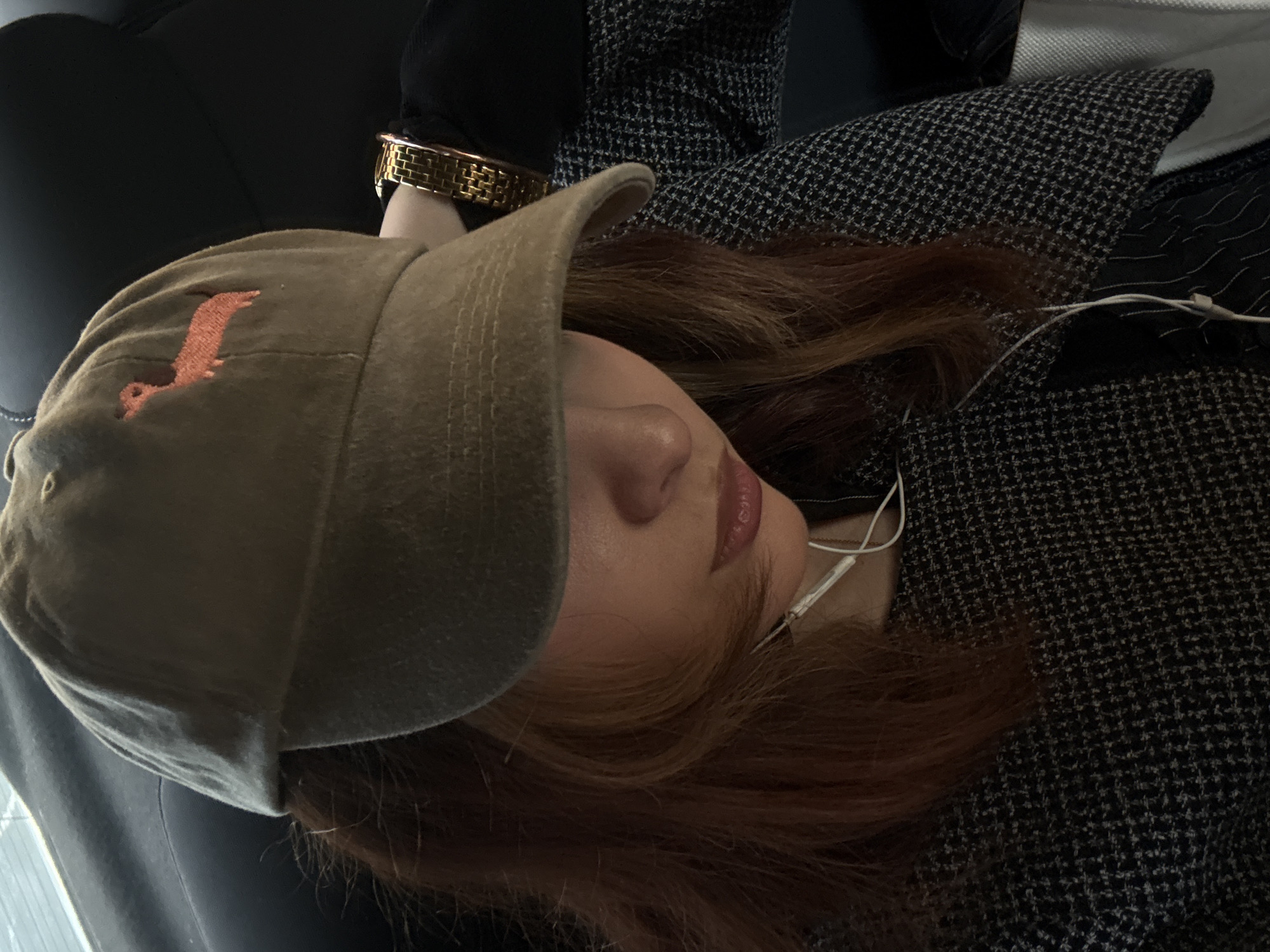 A person with reddish-brown hair is shown from an overhead angle, lying down on what appears to be a dark surface, possibly a car seat. The person is wearing a light brown baseball cap with an orange emblem, a gold-colored bracelet, and earphones. They are also dressed in a black and white patterned garment. The location is unknown, and there is no information to suggest it is Yerevan, Armenia.