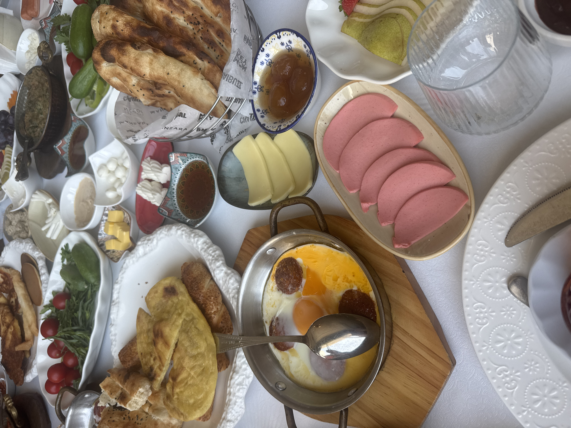 The image depicts a lavish breakfast spread, likely at a hotel or restaurant in Sabail Raion, Azerbaijan. The table is laden with a variety of dishes, including a pan of fried eggs and sausage, sliced cheeses, jams, fresh vegetables like tomatoes and cucumbers, a basket of assorted breads, and a plate of pink sliced sausages. There are also small bowls containing what appear to be olives, yogurt, and a spread. The setting is elegant, with ornate plates and serving dishes. The overall impression is one of abundance and a delightful start to the day.
