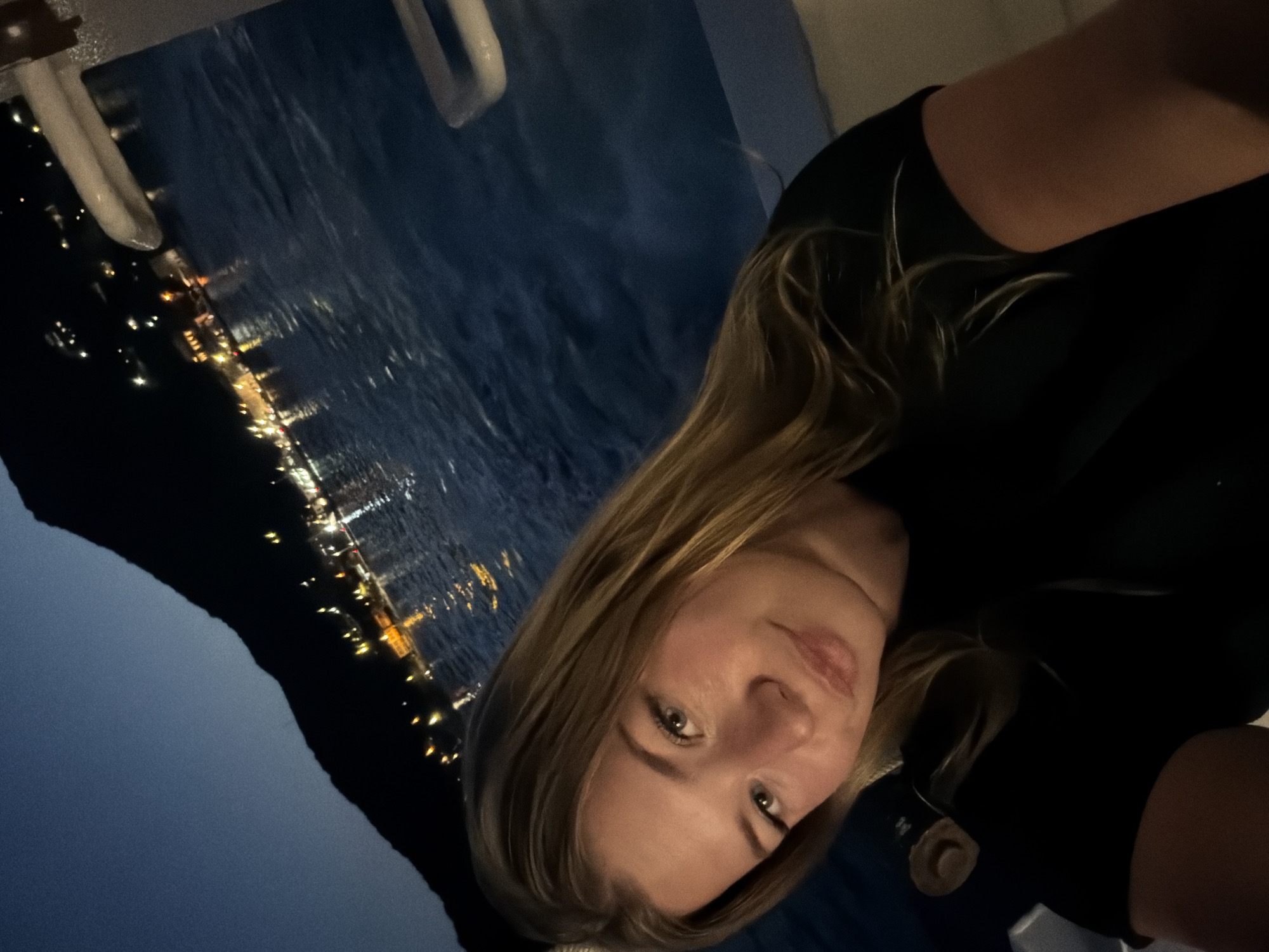 A young woman with blonde hair and light skin takes a selfie at dusk. She is wearing a black shirt. The background shows a body of water, with lights from buildings and mountains visible in the distance. The scene is likely Đurići, Montenegro, given the presence of mountains and proximity to the coast. The lighting suggests it is evening, with the sky a deep blue.