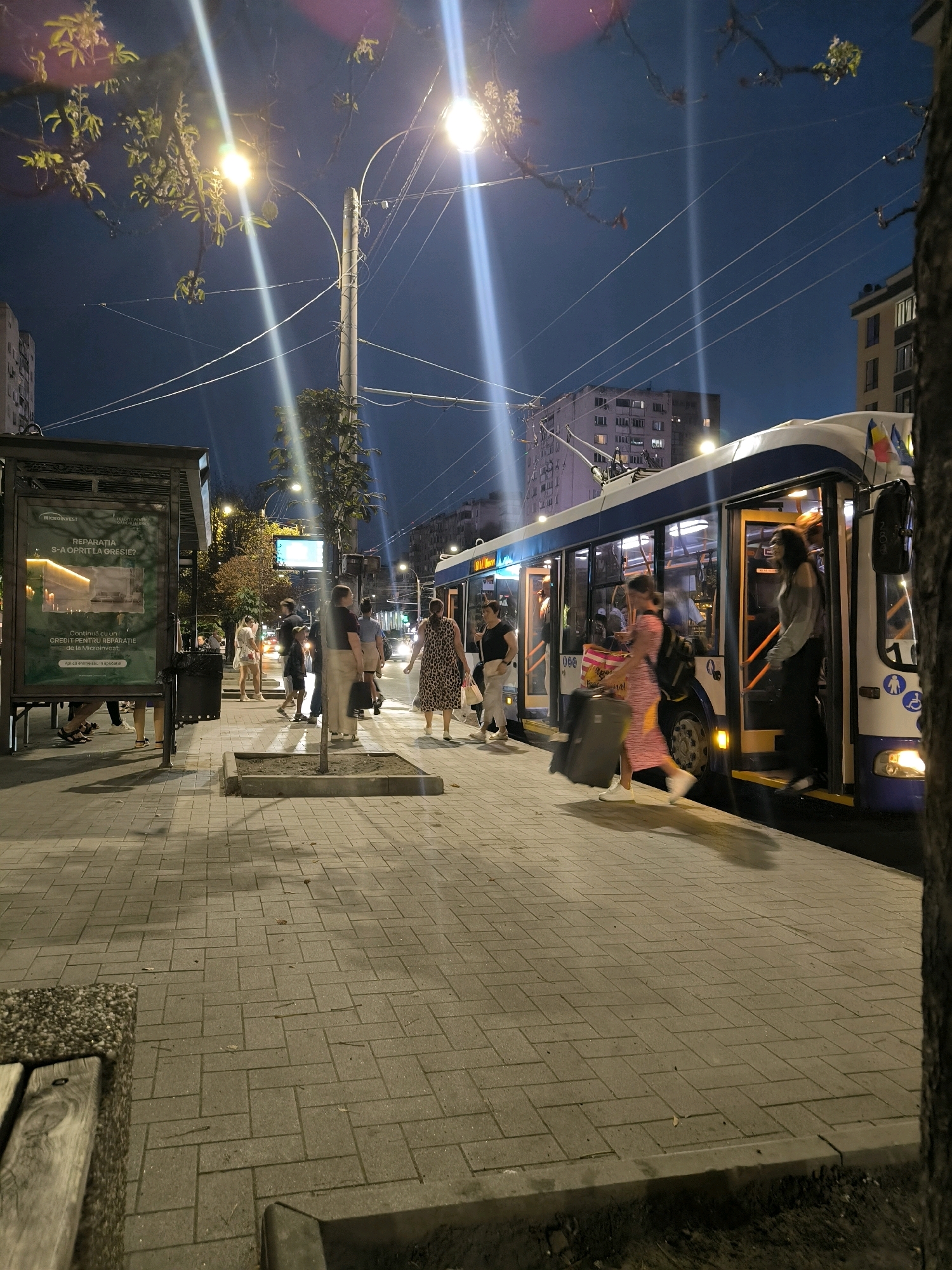 This is a nighttime, eye-level, full shot of a trolleybus stop in Chișinău, Moldova. The trolleybus, numbered 10, is at the stop with its doors open. Several people are either boarding or disembarking, some with luggage. A woman in a patterned dress is exiting the bus, pulling a suitcase. Another woman in a grey top is ascending the steps of the bus. On the sidewalk, several other individuals are waiting or moving around. A bus shelter with an advertisement is visible on the left. The ground is paved with interlocking bricks, and a wooden bench is partially visible in the foreground. Streetlights illuminate the scene, creating long shadows. Overhead, power lines for the trolleybus are visible against the dark blue night sky, with tree branches and leaves hanging into the frame from the upper left. Buildings can be seen in the background. The flags of Romania and Moldova are attached to the front of the trolleybus.