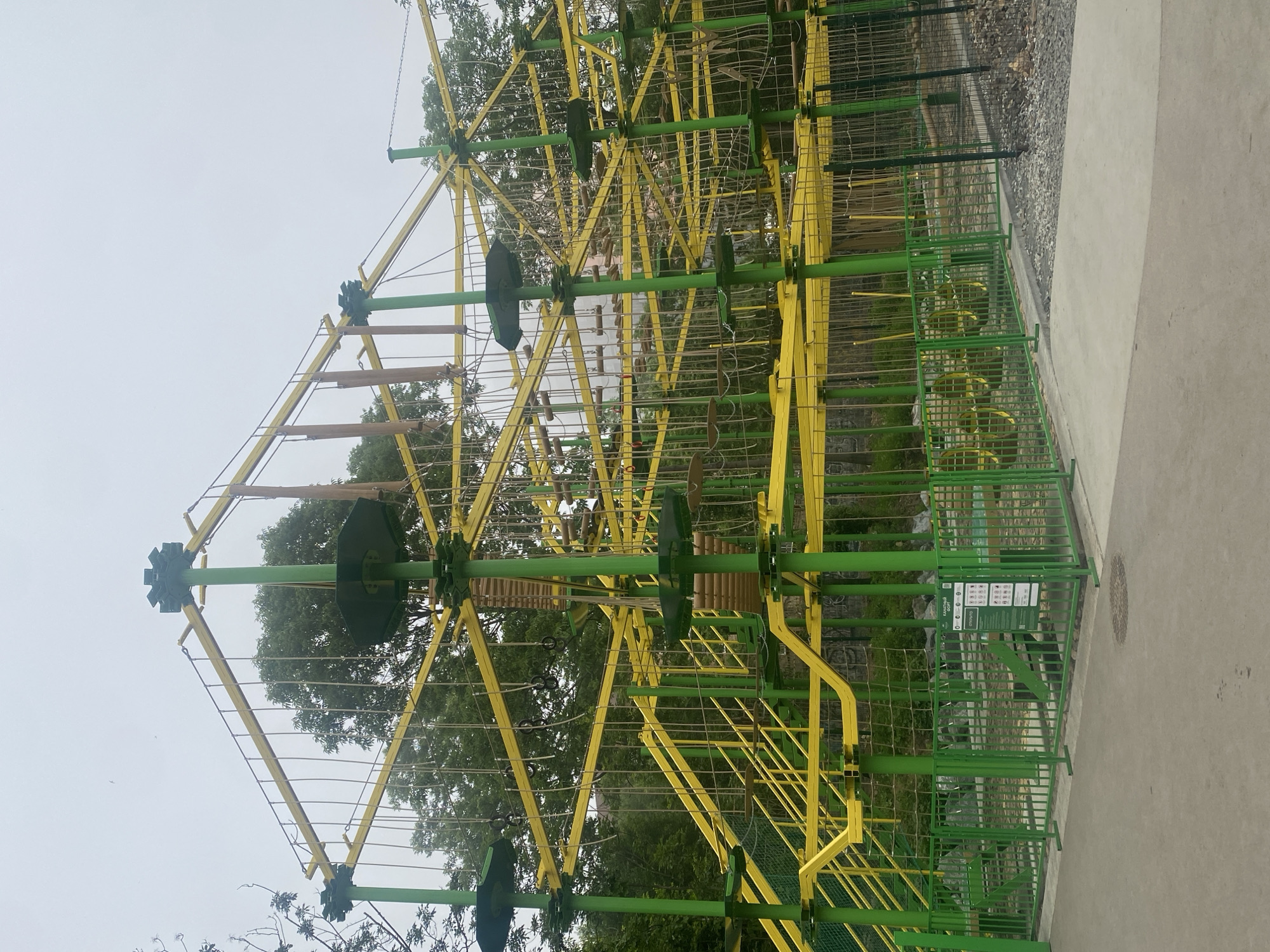 An outdoor adventure rope course is depicted in this image. The structure is primarily green and yellow, with vertical green poles supporting a complex network of horizontal yellow beams. Suspended ropes, wooden obstacles, and platforms are visible at various levels within the structure. A green metal fence encloses the base of the rope course, and a paved walkway runs in front of it. Trees with green foliage surround the structure. The sky is overcast, suggesting a cloudy day. There are no people visible in the image.