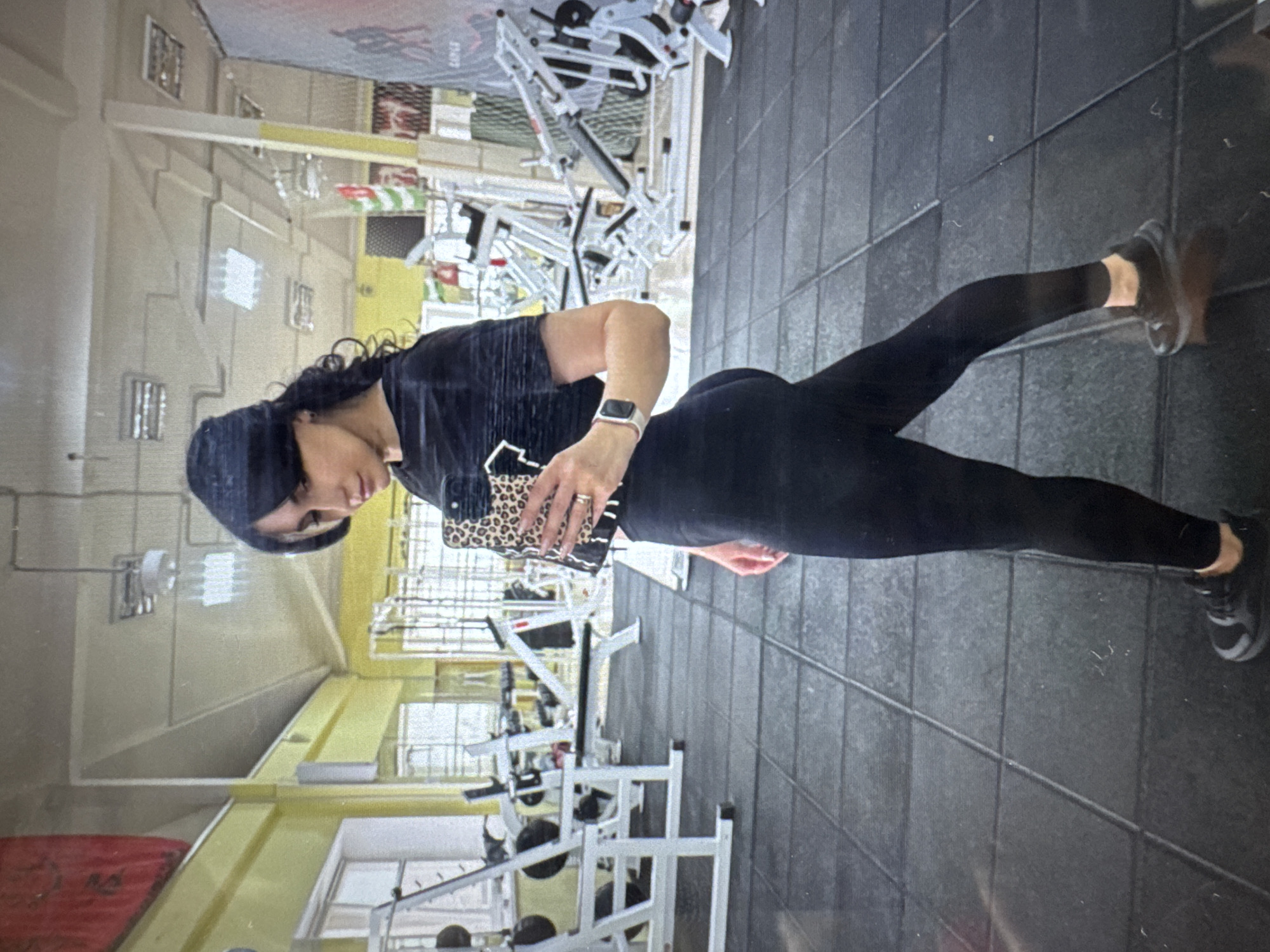 A woman wearing a black t-shirt, black leggings, and a black head covering is standing in a gym in Chaikovsky, Russia, posing for a picture. She is holding a cell phone with a leopard-print case, obscuring her face. She is wearing an Apple watch on her left wrist. Gym equipment is visible in the background. The floor is covered in square, dark-gray tiles.