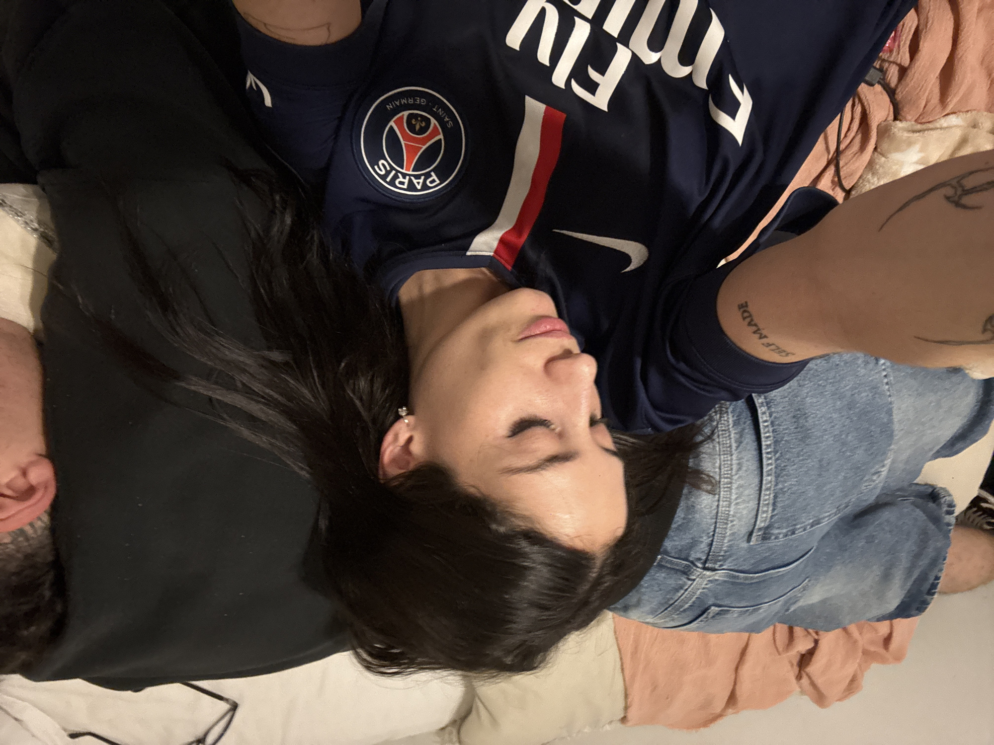 A woman wearing a Paris Saint-Germain jersey, blue jeans, and earrings lies on a bed, next to a person wearing a black shirt and glasses. The location is unknown and cannot be determined from the image.