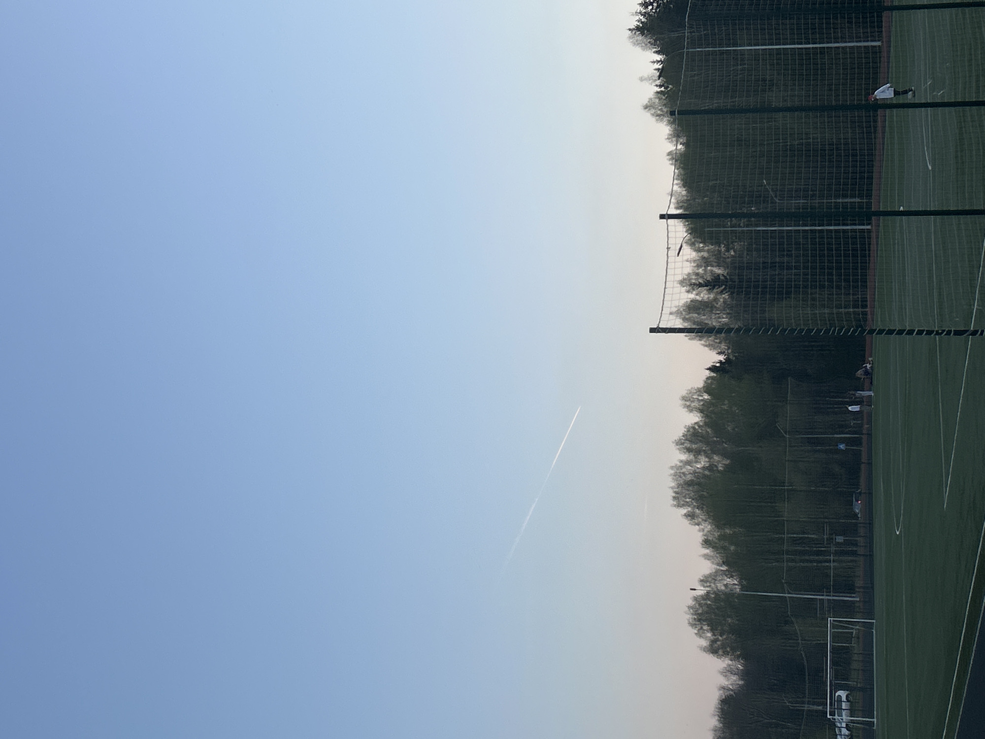 A daytime shot of a sports field in Markovskii, Russia. The sky is blue, with a contrail from an airplane visible. The field is green, with white lines marking the boundaries. A net surrounds the field, and trees are visible beyond the net. A person is present on the field. There is a goal net along the edge of the frame.