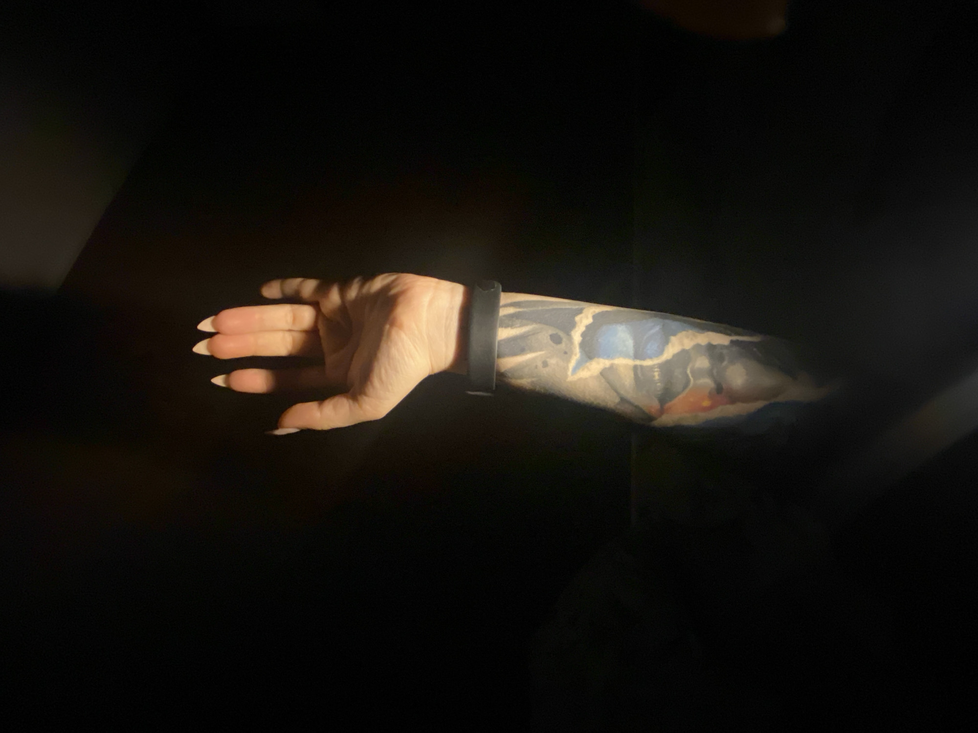The image displays a person's left arm, brightly illuminated by a focused light source, set against an almost entirely dark background. The arm is extended horizontally with the hand slightly open and fingers gently curved. A prominent full-sleeve tattoo covers the forearm and extends up the arm, featuring abstract designs with dominant blue and hints of orange and lighter tones, resembling organic or fluid shapes. A plain, dark-colored band, possibly a watch or fitness tracker, is worn on the wrist. The fingernails are long, light-colored, and appear manicured. The strong, directional lighting creates high contrast, emphasizing the textures of the skin and tattoo, while the surrounding environment remains in deep shadow. No specific activity is depicted beyond the arm being presented for the photograph. No text is visible in the image. The provided location context is Chaikovsky, Russia.