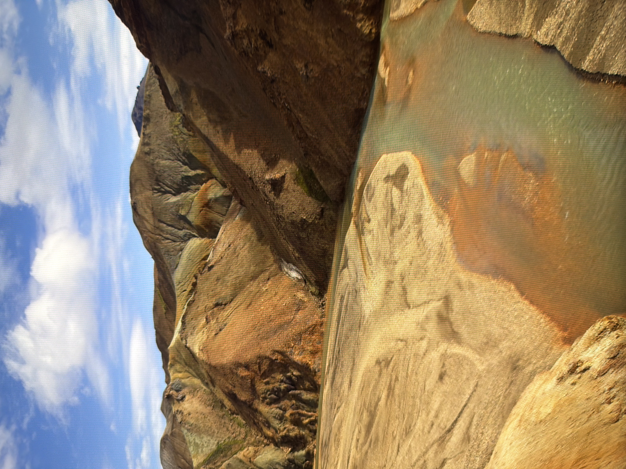 The image depicts a natural landscape with rolling hills, a river, and a cloudy blue sky. The hills are multi-colored, with shades of brown, orange, yellow, and green, suggesting a geologically diverse area. A river flows through the foreground, its water a clear, slightly greenish hue, with sandy banks. The sky is bright blue with fluffy white clouds. The scene appears to be a remote, unpopulated area. There is no indication of the city of Kyiv, Ukraine, or any urban elements.