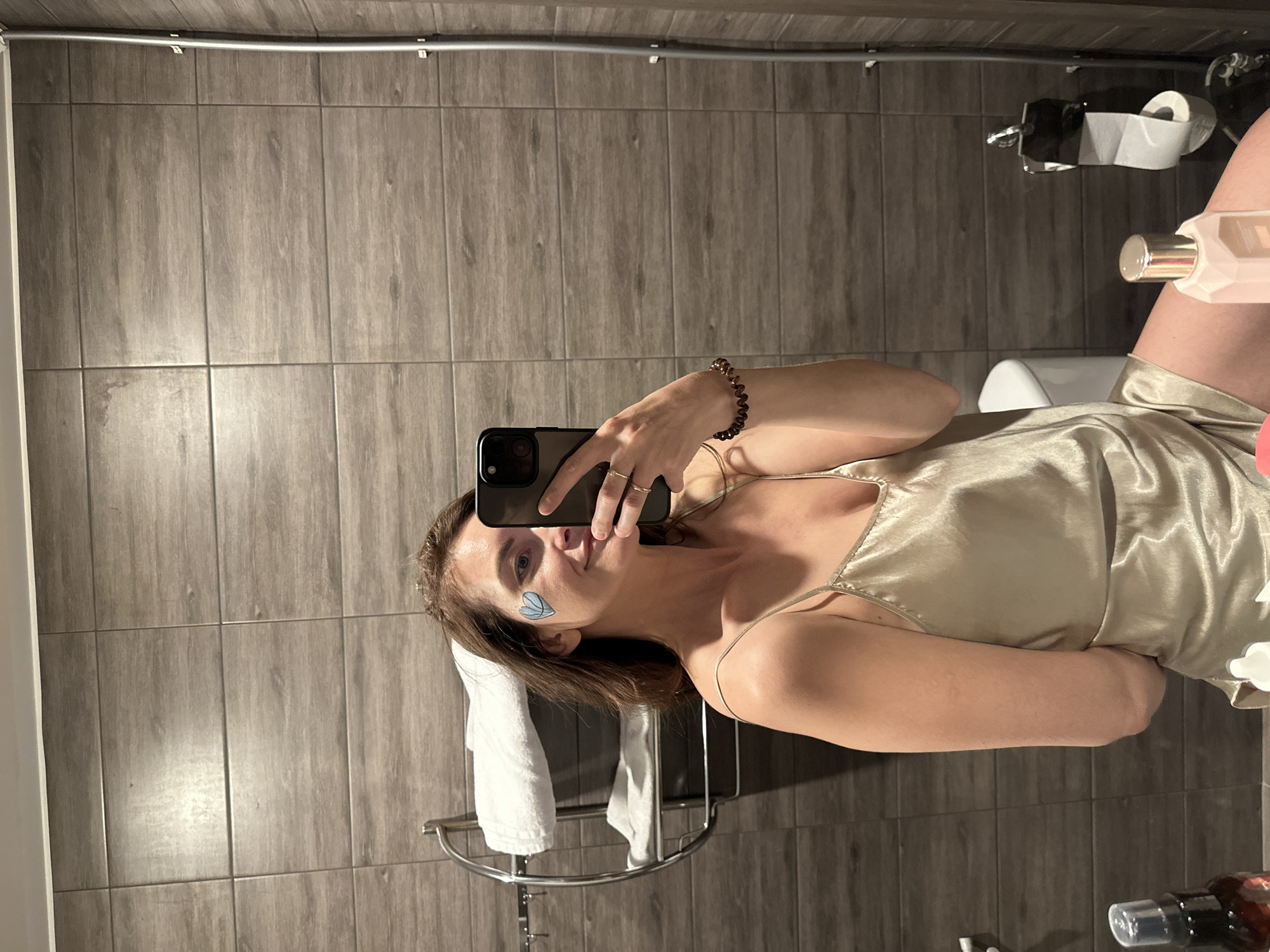 A woman stands in a bathroom, taking a selfie with her smartphone. The bathroom has gray tiled walls. The woman is wearing a silky, light gold-colored top with spaghetti straps. She has a small, blue, butterfly-shaped sticker on her cheek. She is wearing a bracelet on her right wrist. Behind her is a towel rack with a white towel hanging on it. In the background, there is a toilet, a roll of toilet paper, and various bottles on the floor. The specific location in Saint Petersburg, Russia cannot be determined from the image.