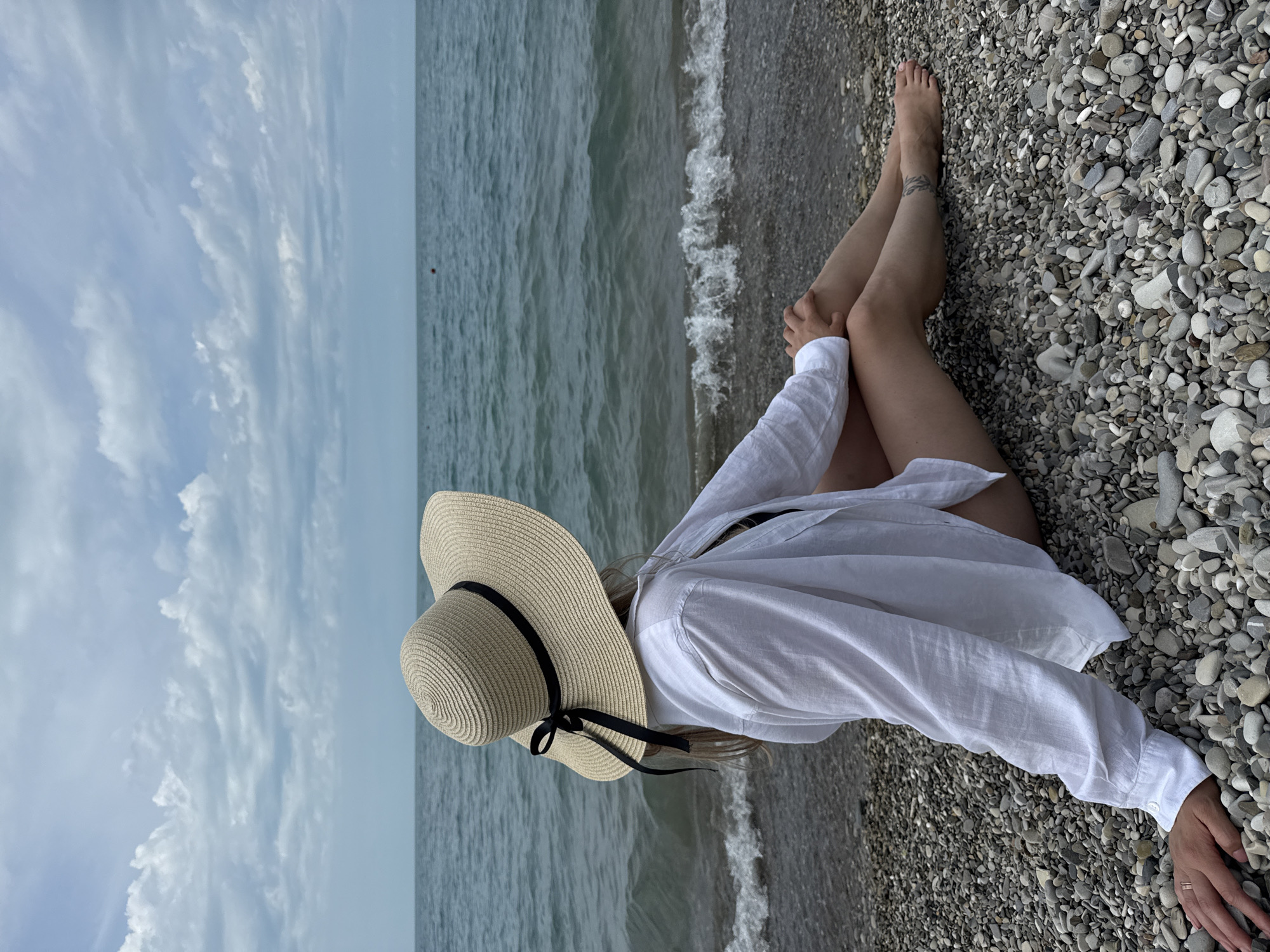 A woman is sitting on a pebble beach in Nebug, Russia, looking out at the sea. She is wearing a large straw hat with a black ribbon, a white open-buttoned shirt, and what appears to be a black bikini underneath. Her legs are crossed, and her arms are resting on her knees. There is a small tattoo visible on her right ankle. The sea is calm with small waves lapping at the shore. The sky is overcast with scattered clouds. The beach is composed of small, grey and white pebbles.