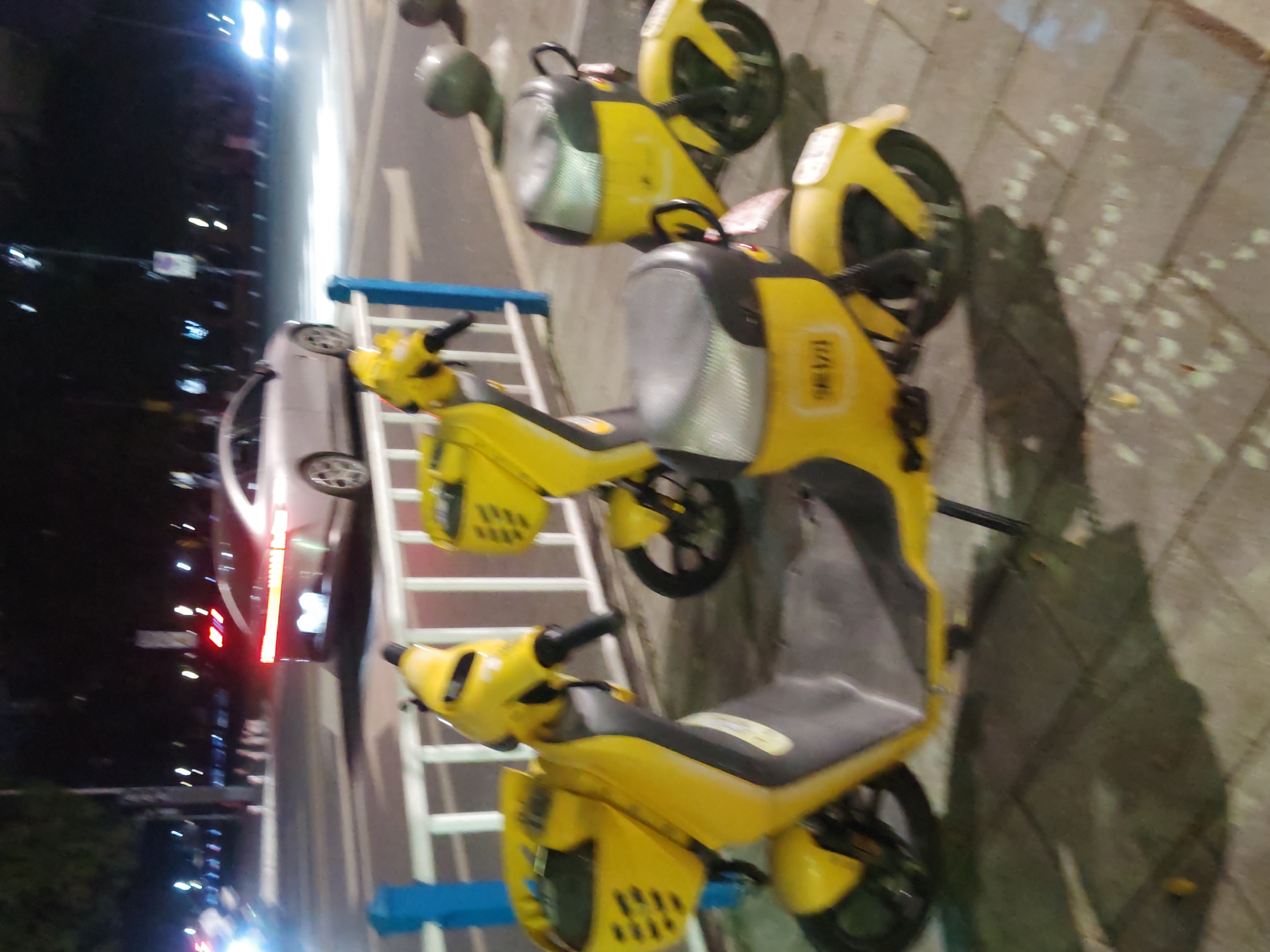The image shows a row of parked yellow electric scooters along a sidewalk in Chongqing High-tech Zone, China. A white car is parked next to the scooters. A white and blue ladder-like structure is between the car and the scooters. The scene is at night.