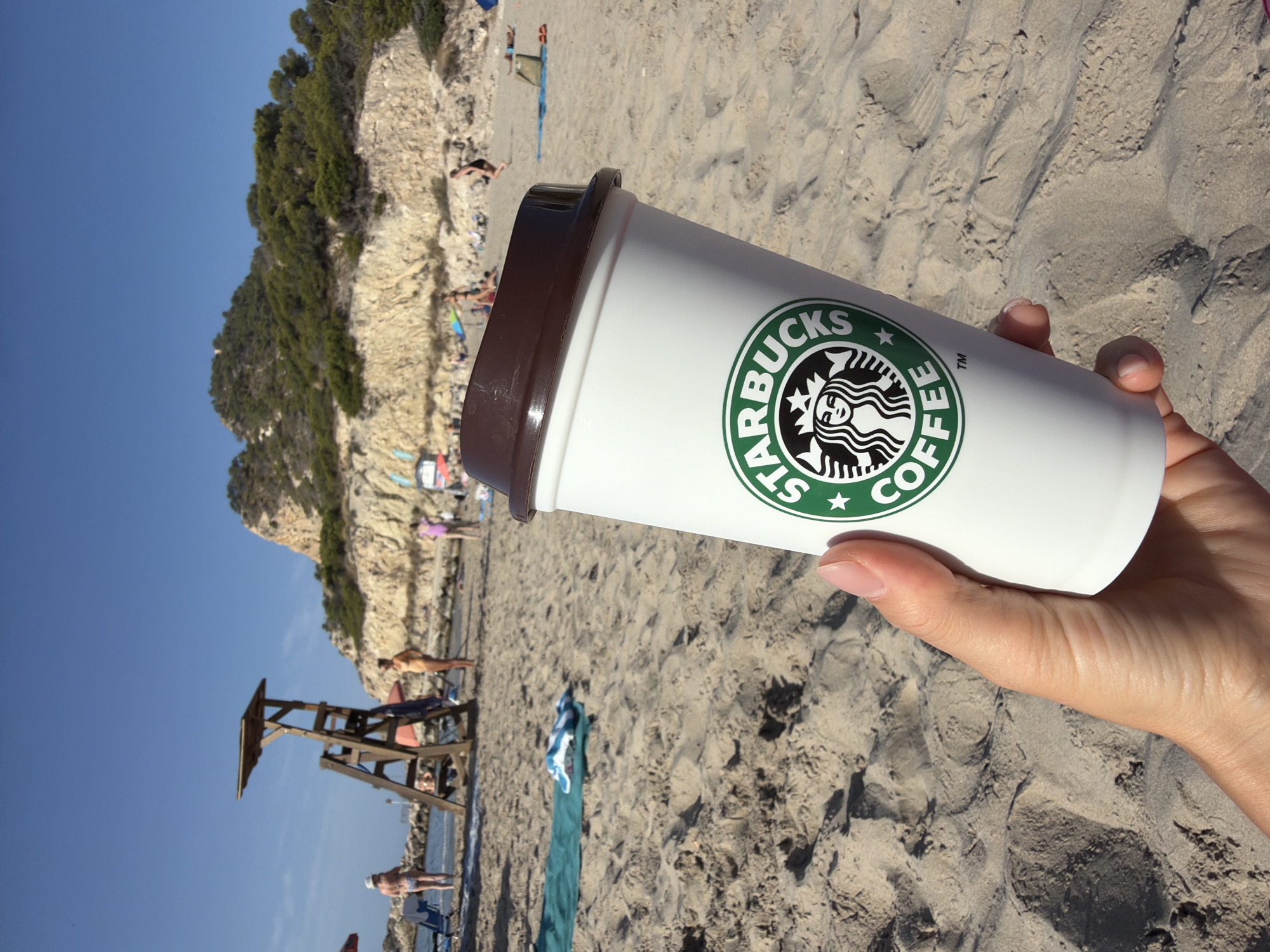 A hand holds a white Starbucks coffee cup with a brown lid in the foreground. The cup has the Starbucks logo printed in green and white. The background depicts a sandy beach under a clear blue sky. Several people are visible on the beach, some near the water and others spread out on the sand. A wooden lifeguard stand is situated further back on the beach, and a rocky, tree-covered hill rises behind it. The scene appears to be set in Urbanització Cales I Talaies, Spain, based on the visual cues of a Mediterranean beach environment.