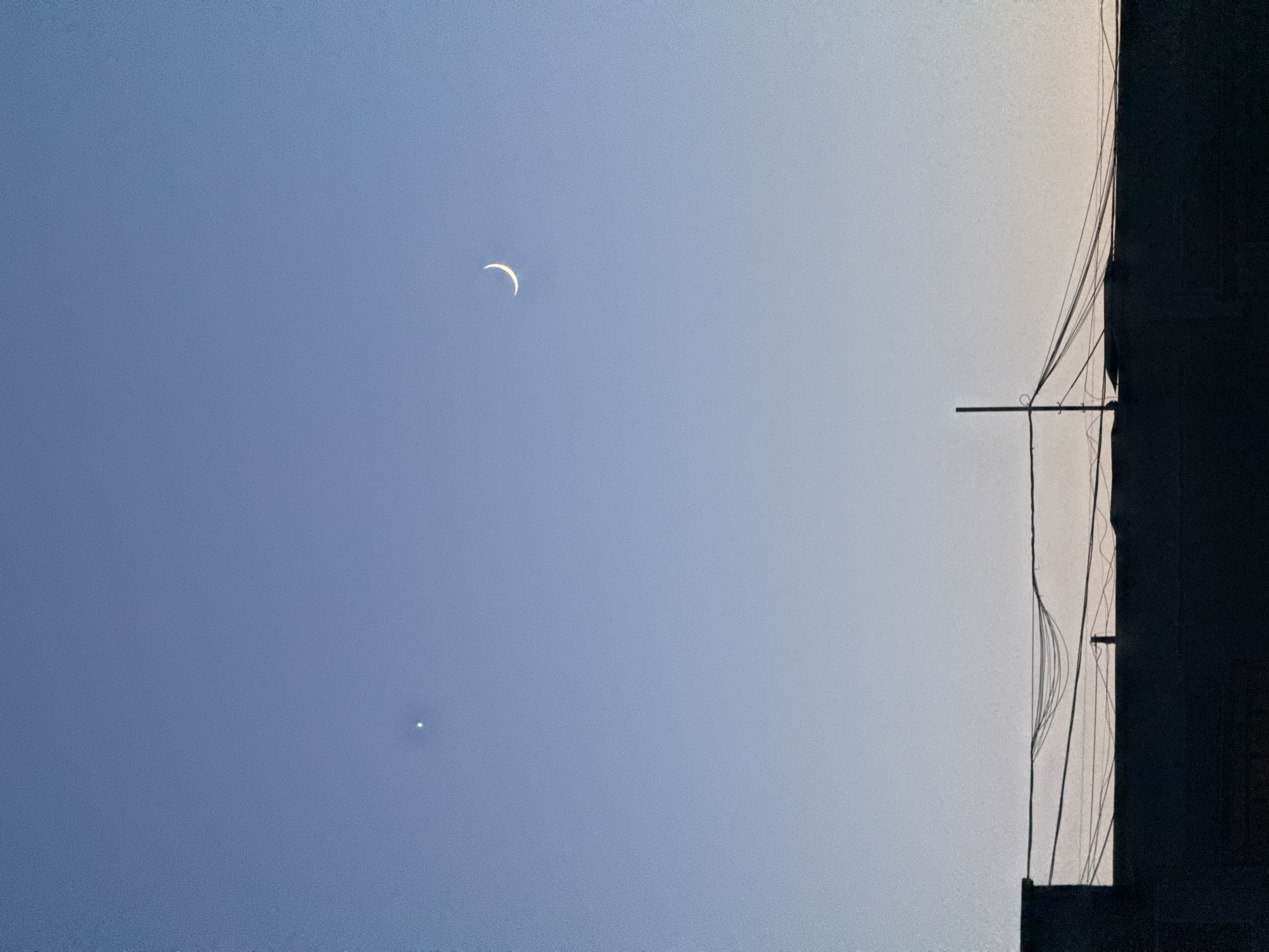 The image shows a crescent moon in a blue sky, with a bright star or planet visible below it. To the right, there is a silhouette of a building with wires running down its side. The image was taken in the city of Kazan, Russia.