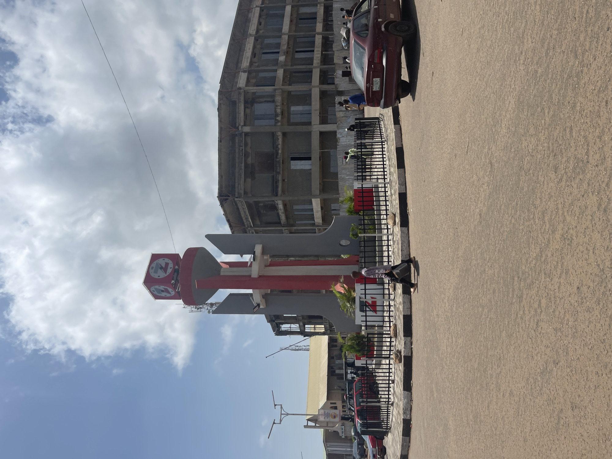 The image shows a street scene in Jos, Nigeria, featuring a prominent clock tower monument in the foreground. The monument is red and grey, with four clock faces visible at its apex. The monument is surrounded by a black metal fence with a white and black striped border. Several cars are parked or driving in the vicinity. Behind the monument, a large, multi-story building under construction is visible, with visible scaffolding and unfinished concrete structure. To the left of the monument, a more completed building with a yellow roof and church-like steeple is visible. The sky is partly cloudy, with bright sunlight indicating daytime. A single person is walking across the foreground, carrying a dark-colored bag. Other individuals are present near the construction site and on the sidewalk. The road surface is asphalt.