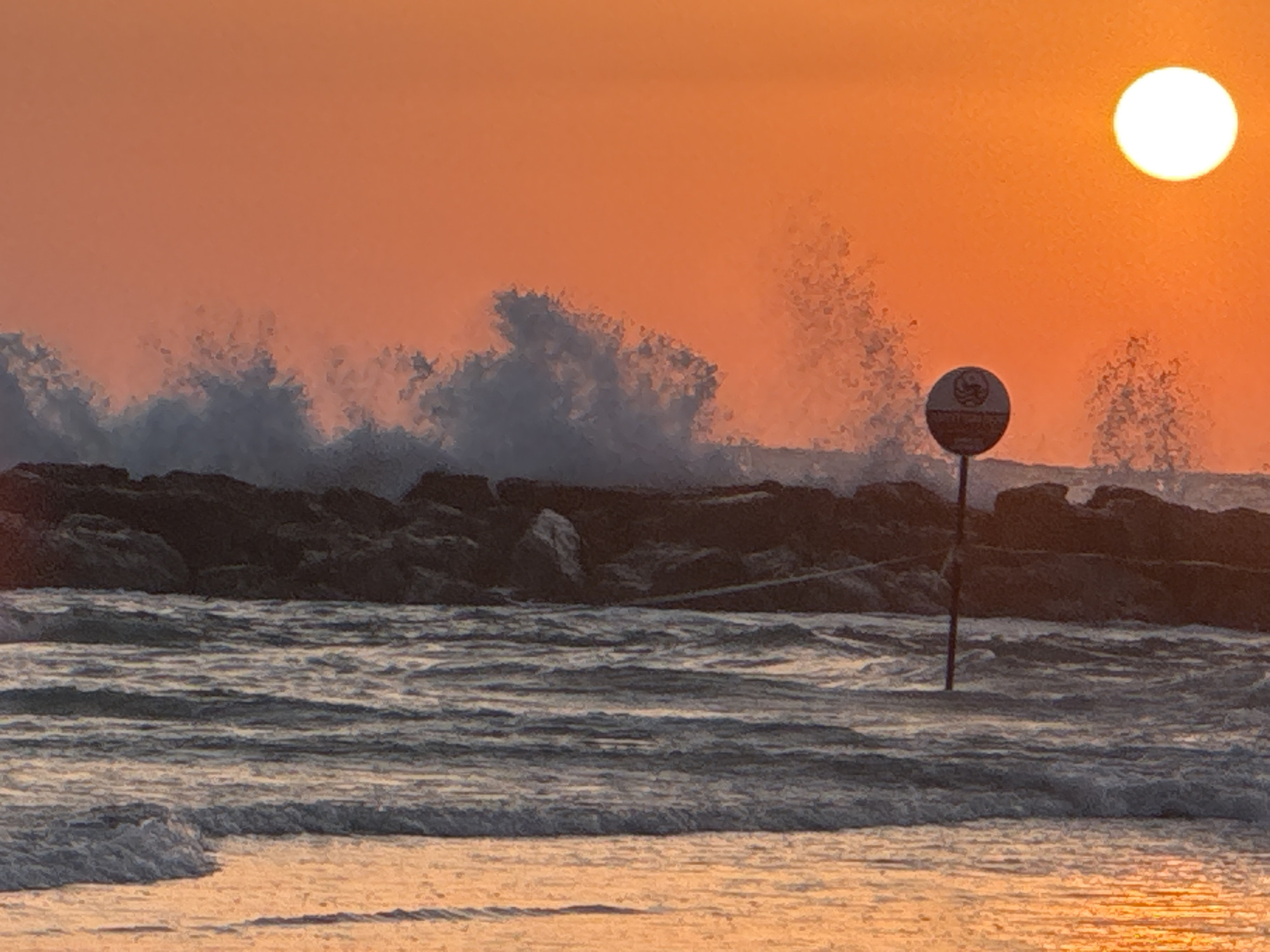 The image captures a sunset over the sea in Netanya, Israel, likely at Herzl Beach given the presence of a distinctive circular sign on a pole in the water. The sky is a vibrant gradient of orange and pink, with the sun a bright white orb in the upper right corner. Waves are crashing against a rocky breakwater, creating white spray against the dark rocks and the colorful sky. The foreground shows the gentle lapping of smaller waves on a sandy beach, illuminated by the warm sunset light. The sign, featuring a circular logo with Hebrew text and imagery, is a key identifier of the location. No people are visible in the scene.