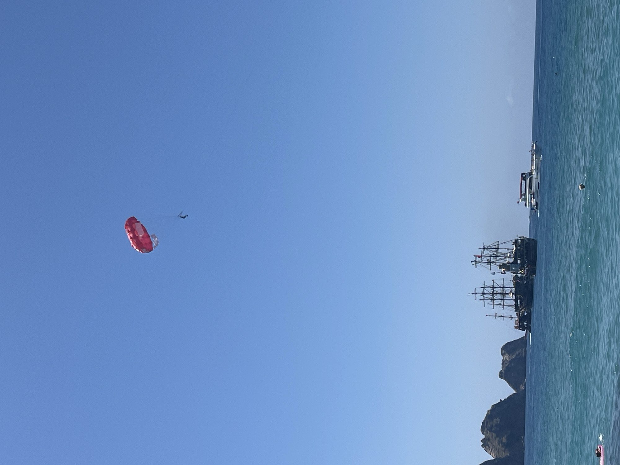 The image captures a coastal scene in Alanya, Turkey. A blue sky dominates the view, with a red parasail and two unidentifiable figures suspended mid-air to the left. To the right, the turquoise sea is visible. There are three boats on the right side of the image: one appears to be a modern vessel, and the other two are traditional tall sailing ships, reminiscent of pirate ships. Rocky land formations can be seen along the coast.