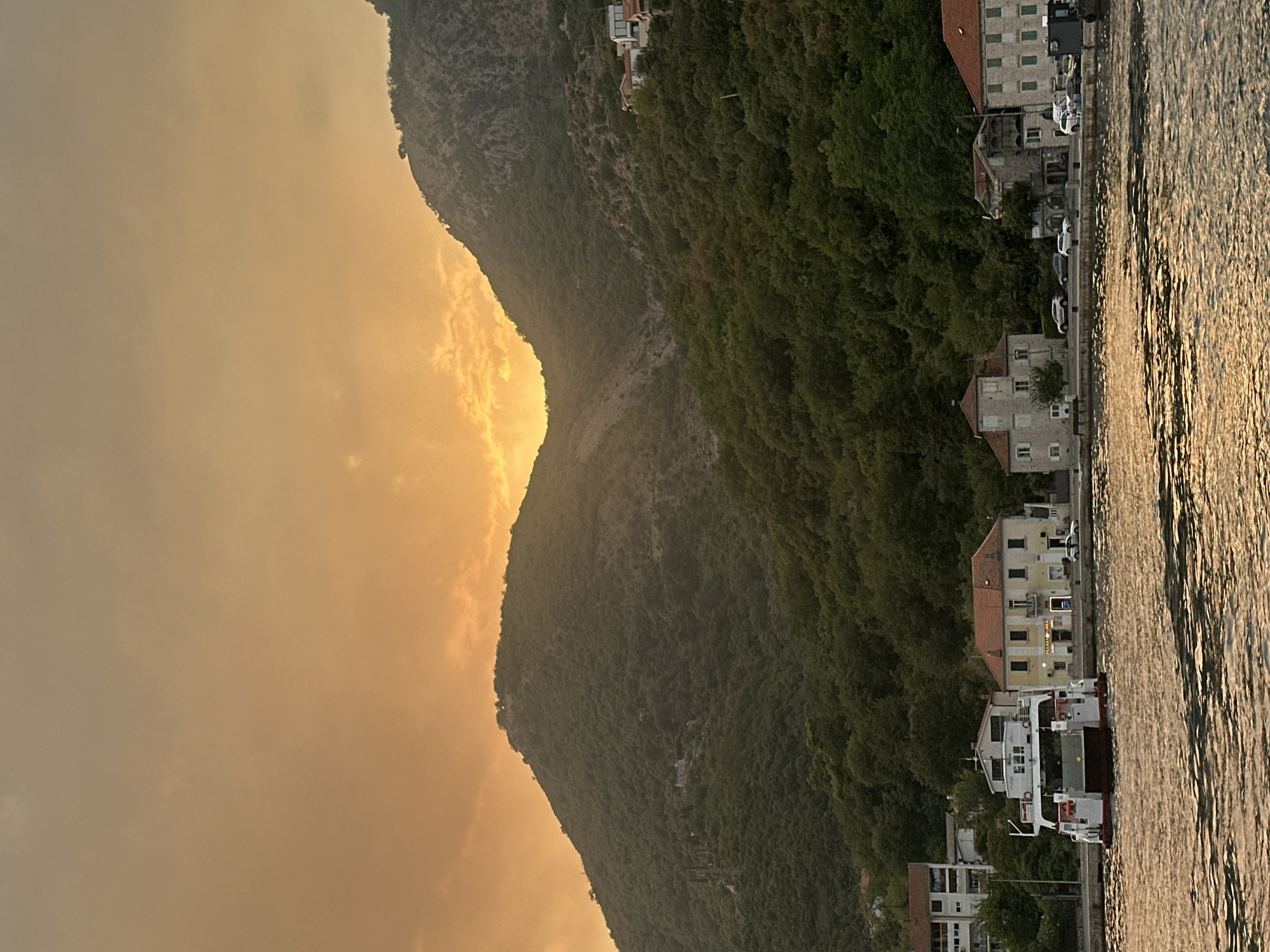 The image shows a sunset over a bay in Jošice, Montenegro. The sky is a warm orange and yellow, with some clouds. A steep, tree-covered mountain dominates the background. In the foreground, there is a body of water with ripples reflecting the sunset light. Along the shore, there are several buildings, including a ferry docked near a yellow building with a sign that reads "PIZZERIA LUNA". There are also some cars parked along the waterfront and a few people visible near the buildings. The scene appears to be peaceful and serene.
