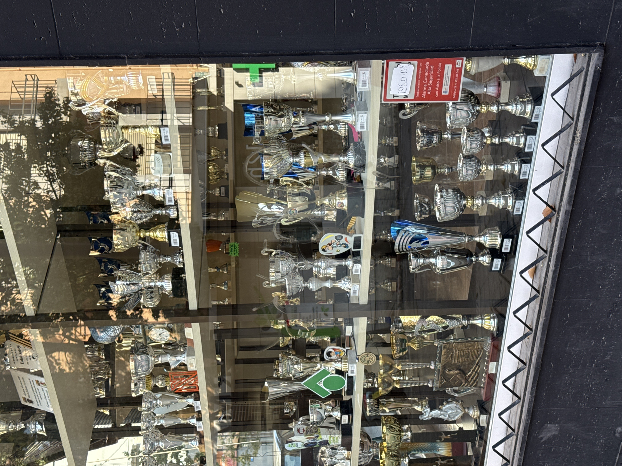 A vertical shot captures a shop window filled with numerous trophies, reflecting the exterior environment. The window displays dozens of trophies of various sizes, shapes, and materials, primarily gold-toned and silver-toned, with some featuring blue accents or clear plastic/glass components. Many trophies depict generic athletic figures, while notable ones include a Mickey Mouse figurine, a trophy with a clock, and one featuring a green baseball field graphic with "LO" or "10" on it.
Strong reflections on the glass obscure parts of the display, showing trees, buildings, and what appears to be a sunny outdoor scene, suggesting it is daytime. The shop window is framed by a dark, possibly black, border at the top and a decorative silver-grey frame with a zigzag pattern on the right side.
A prominent red sign with white text is visible in the upper right portion of the window. The text reads: "NIOSDINA", "Estudiantes", "Establecimiento protegido con: Alarma Conectada Alta Seguridad con Aviso a la Policía", and "Teléfono 24 H: 902 366 366". Small, unreadable labels are attached to some trophies.