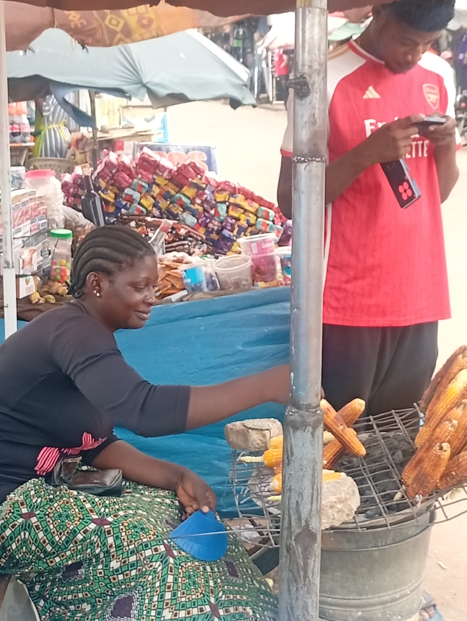 A woman in Sabon Tasha, Nigeria, is grilling corn on a charcoal grill at her outdoor vending stand. She is sitting, wearing a dark long-sleeved shirt and a patterned green, white, and brown skirt. A blue fan sits in her lap. To her right is a metal charcoal grill topped with corn. Behind her are stacked packages of goods. To her right, a young man in a red and white soccer jersey is looking at his cell phone. He is standing near the grill. A metal support pole is in the foreground, partially obscuring the scene. An umbrella provides shade. In the background are other people and structures indicating an outdoor market setting.