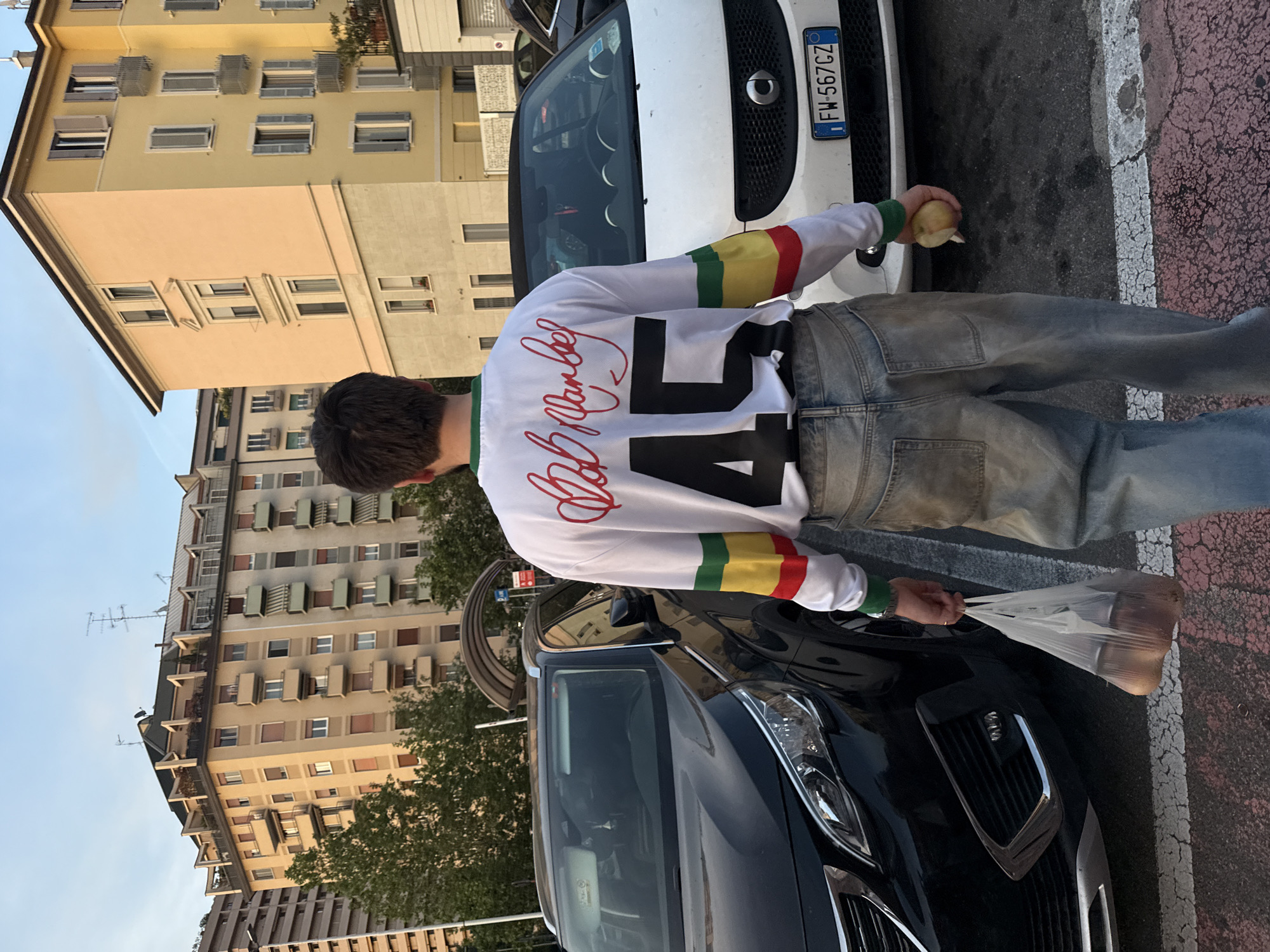 A medium shot from behind of a male in jeans and a white long-sleeve shirt with green, yellow, and red striped accents on the arms and a large black "45" on the back, with red script above the number. He is holding a single yellow pear in his right hand and a clear plastic bag filled with similar fruit in his left hand.
He is standing between a white Smart car and a black Peugeot car parked on a street with red and white striped road markings. The license plate of the Smart car, "FW-567CZ," indicates the location is Milan, Italy.
In the background, there are several multi-story buildings with apartments, and trees along the street. The sky is light blue.