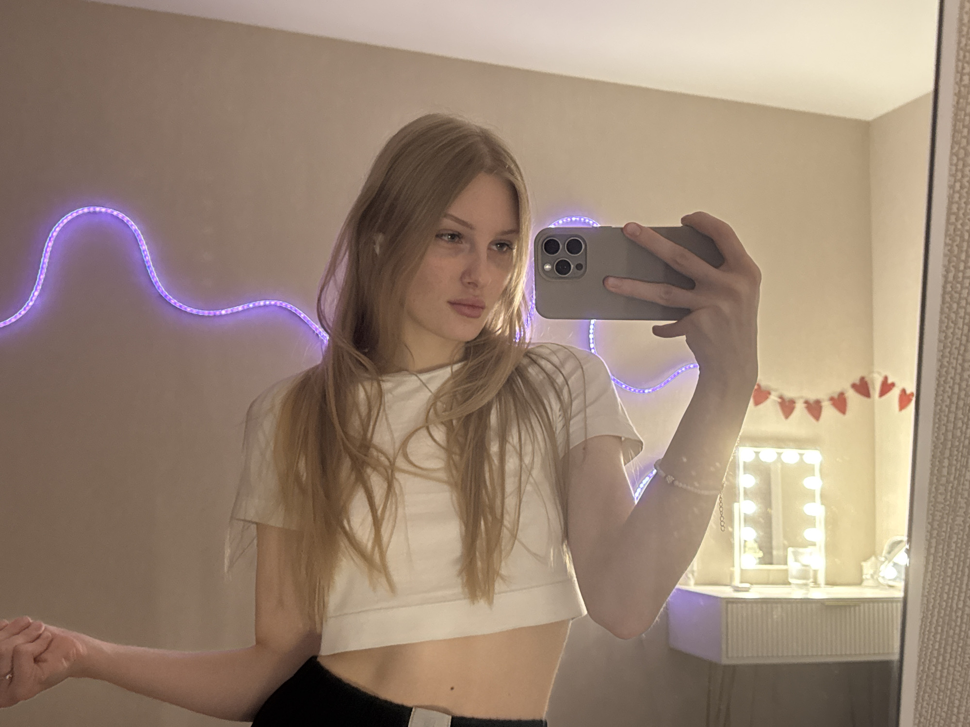 A young woman is captured taking a mirror selfie indoors. She has long, straight, light brown hair and is wearing a white cropped t-shirt over dark bottoms. She holds a grey smartphone, likely an iPhone due to its distinctive multi-lens camera, with her right hand, positioned to reflect her face and upper body. A white earbud is visible in her left ear. Her expression is neutral, with her gaze directed slightly to her right.
The background features a light, neutral-colored wall, against which a wavy purple LED strip light is mounted, casting a soft glow. In the reflection to the right of the frame, a white vanity table with a mirror framed by several circular lights is visible, suggesting a dressing area. Above the vanity mirror, a garland of small red heart shapes is strung across the wall. The woman also wears a light-colored bracelet on her right wrist and a ring on her left hand. The lighting from the LED strip and vanity lights indicates an indoor setting, likely in the evening or night. There is no visible text.