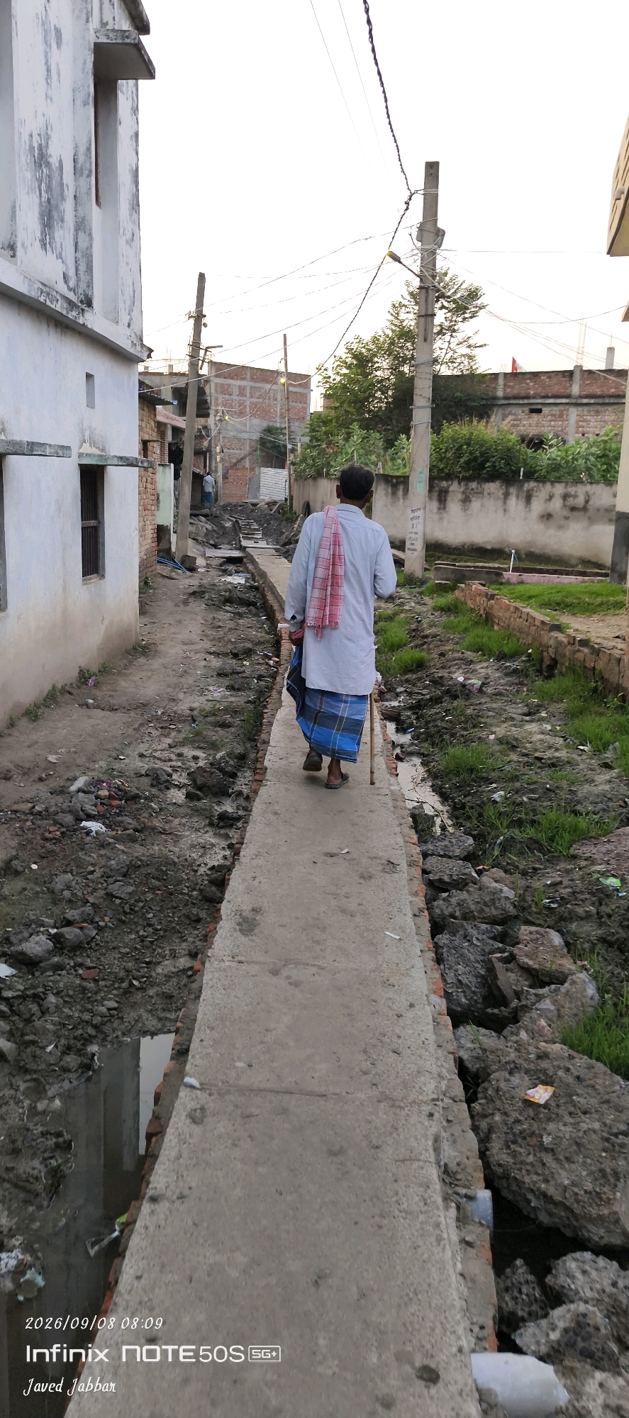 A man with a pink and white checkered cloth draped over his shoulders walks down a narrow concrete path in a village in Bahera, India. He is wearing a light-colored shirt and a blue and white checkered lungi. The man is using a wooden cane for support. The path is flanked by a dirty, overgrown drainage ditch on one side and a rough brick wall and a white building on the other. The environment appears to be an urban or semi-urban area with visible construction and overhead power lines. The ground is littered with debris and some patches of green grass are visible. The sky is overcast, suggesting it is either early morning or late afternoon.