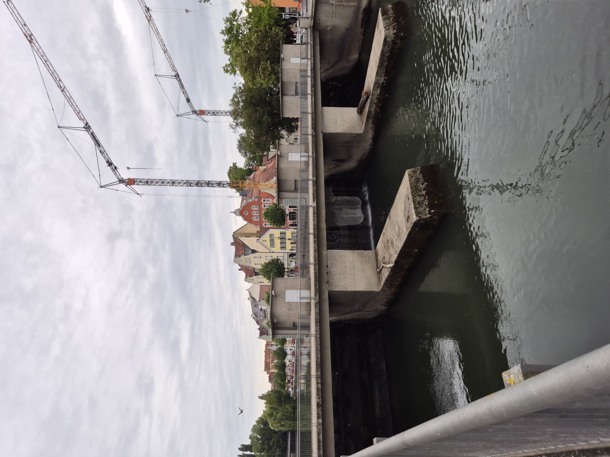 This image depicts a view of the Isar river in Landshut, Germany, with a bridge structure and two construction cranes in the background. The bridge appears to be a pedestrian bridge with railings and a concrete walkway. Beneath the bridge, there are channels of water flowing through concrete structures. In the middle of the bridge, there are two small, grey buildings. Beyond the bridge, a row of colorful buildings lines the riverbank, with trees and lush greenery visible. The sky is overcast with a light gray hue. The scene suggests ongoing construction or renovation work in the vicinity, as indicated by the presence of the cranes. No individuals are clearly visible in the image.
