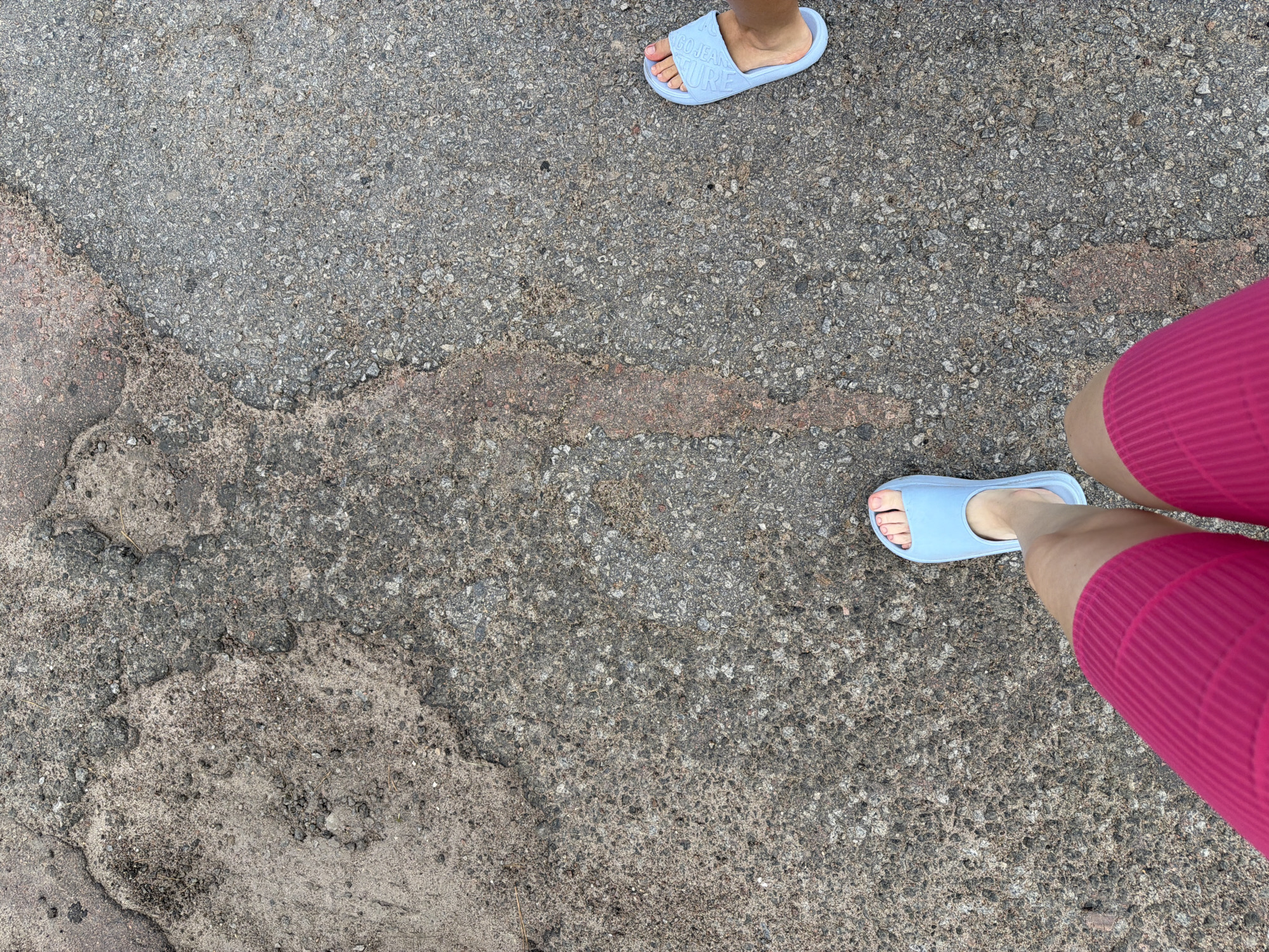 The image depicts a ground-level view of two individuals standing on a textured, asphalt-like surface. The ground shows signs of wear and damage, with patches of lighter-colored material and visible gravel. One individual is wearing bright pink shorts and light blue slides, with their left foot positioned closer to the foreground. The other individual's right foot, also in a light blue slide, is visible in the upper right portion of the frame. The environment appears to be an outdoor urban setting, possibly a road or pathway in Mali Kosharyshcha, Ukraine, given the context. The lighting suggests it is daytime.