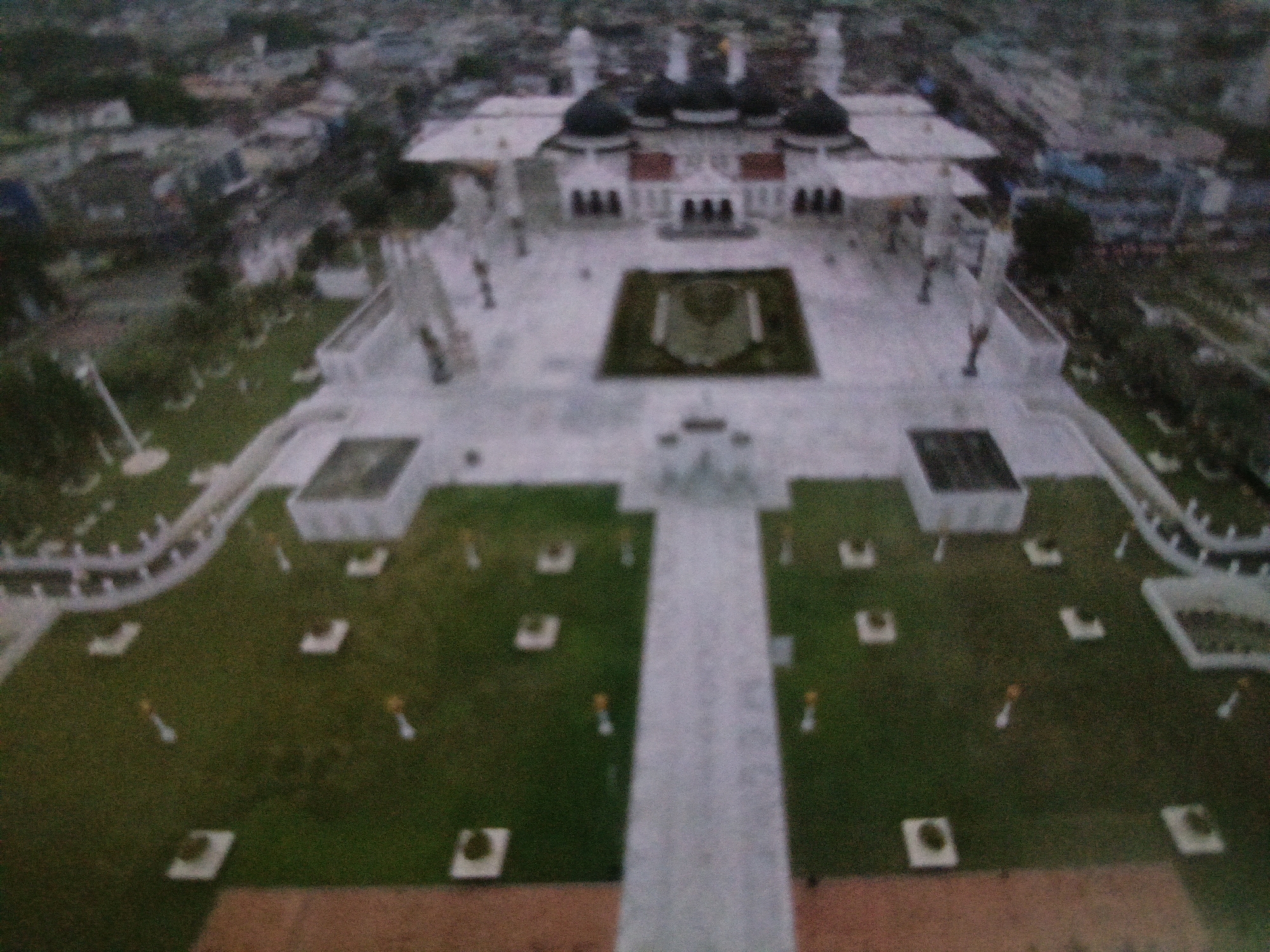 This is a blurred, aerial view of a large, domed building in Gampong Blang, Indonesia. The building appears to be a mosque, with multiple domes and a white facade. In front of the mosque is a large courtyard with a central rectangular garden area. The courtyard is surrounded by manicured grass lawns, which are dotted with small square structures and lampposts. A paved pathway leads from the foreground towards the mosque. In the background, the cityscape of Gampong Blang is visible, with various buildings and trees. The image is taken from a high vantage point, suggesting it was captured by a drone or from a tall building. The overall impression is of a spacious and well-maintained religious complex.
