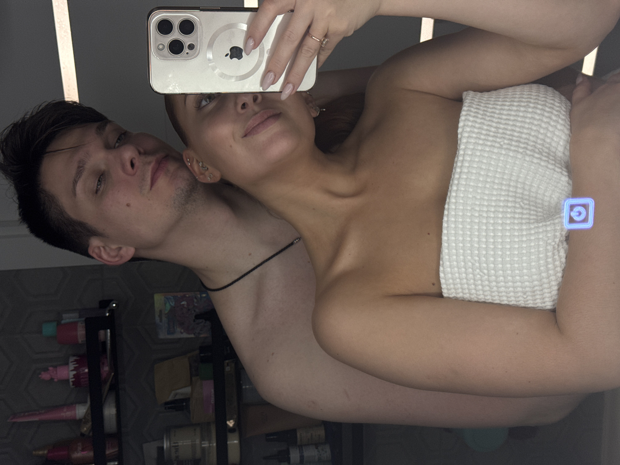 The image depicts a mirror selfie of a man and a woman in what appears to be a bathroom. The precise location within the city of Chaikovsky, Russia, cannot be determined from the visual evidence.
The woman is in the foreground, taking the photograph using a light-colored iPhone with a triple camera system and a circular white accessory (possibly an Apple AirTag or MagSafe puck) visible on its back. She is wearing a white, textured strapless towel, covering her torso. Her head is tilted, and she exhibits a slight smile. She has multiple piercings on her left ear and light-colored nail polish on her fingernails. A ring is visible on the ring finger of her left hand.
Standing directly behind the woman, the man is bare-chested and wears a thin black beaded necklace. He has short, dark hair and a neutral expression, looking towards the mirror reflection. His left arm is positioned around the woman's upper back/shoulder area.
The background, reflected in the mirror, shows a wall with a dark, geometric hexagonal pattern. Two black wall-mounted shelves are visible, holding various toiletries and personal care items, including bottles, tubes, jars, and a small pink decorative figure on the top shelf. A packaged item is on the lower shelf. A blue/purple square icon with a power symbol is overlaid on the lower center of the image.