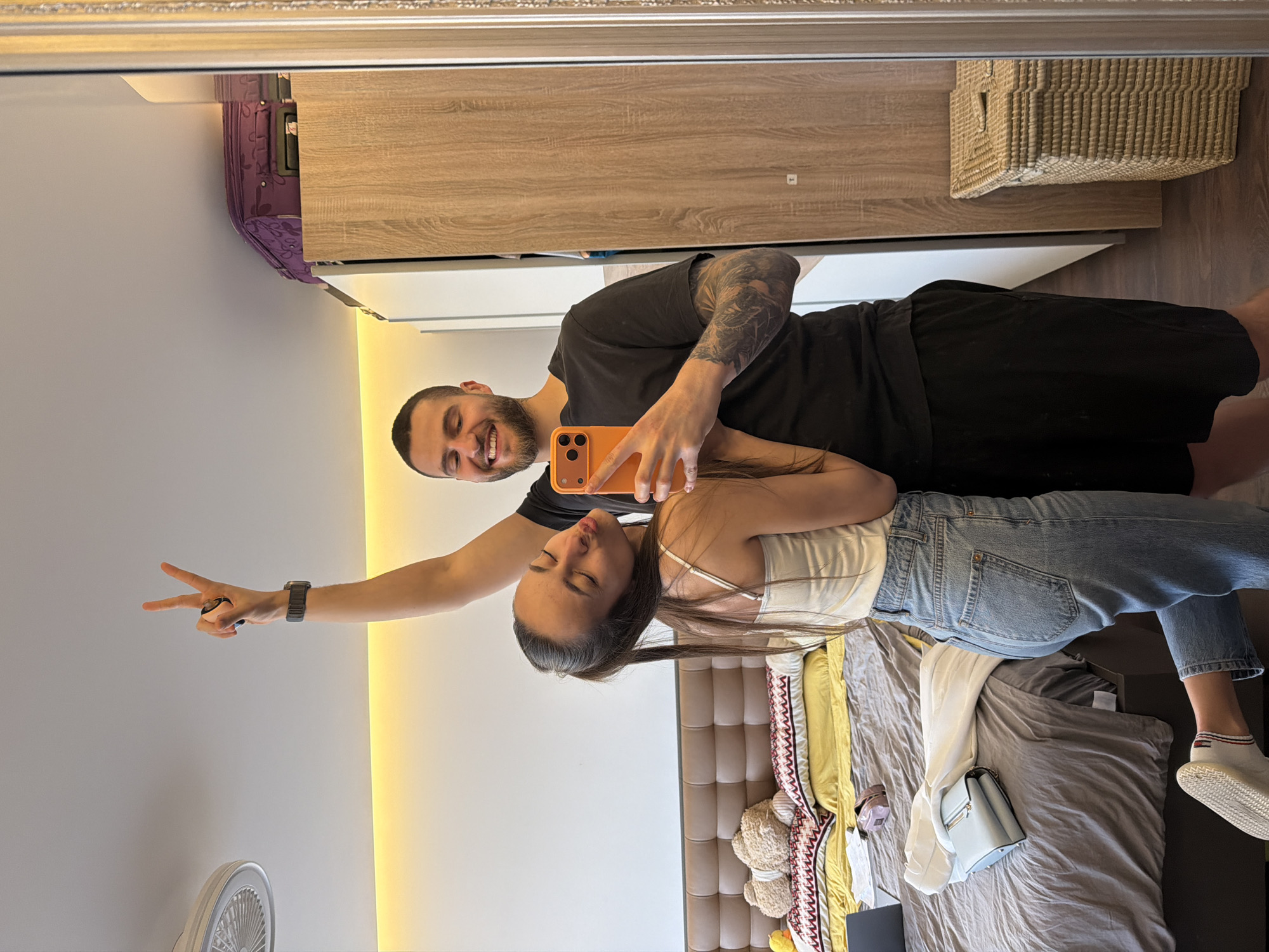 A mirrored selfie features a man and a woman in what appears to be a bedroom. The man, with short hair, a beard, and tattoos on his arm, holds an orange smartphone to take the photo. He is wearing a black t-shirt and black shorts. He is smiling. The woman, with long dark hair and wearing a white top and blue jeans, is positioned close to him. She is pouting, and has closed her eyes. The man extends his other arm, making a peace sign.
The background includes a bed with gray and yellow bedding, a gray headboard, and various objects like a handbag and stuffed animals scattered on the bed. The room also features a wardrobe with light wood and white panels, and a woven storage basket on top. The bottom of a white fan is visible.
The location is not discernible from the image, and therefore cannot be said to be in the city of Sofia, Bulgaria.