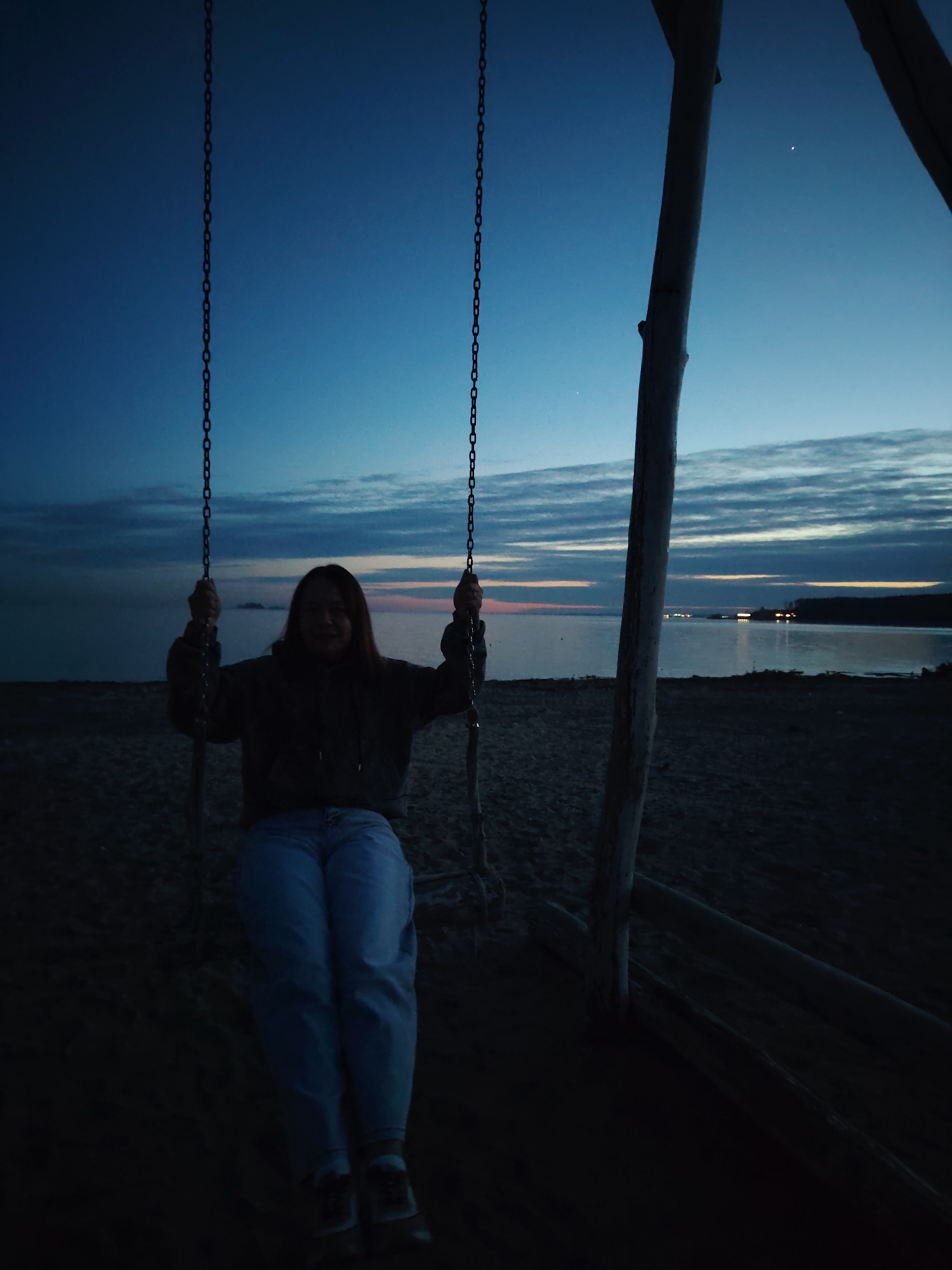 A woman sits on a swing made of rope and chains, hanging from a wooden structure on a beach. The sky is dusk, with a mix of blue and orange hues near the horizon, reflecting on the water in the background. A landmass with distant lights is visible across the water. The beach appears sandy. The woman wears a dark hooded top and light-colored jeans.