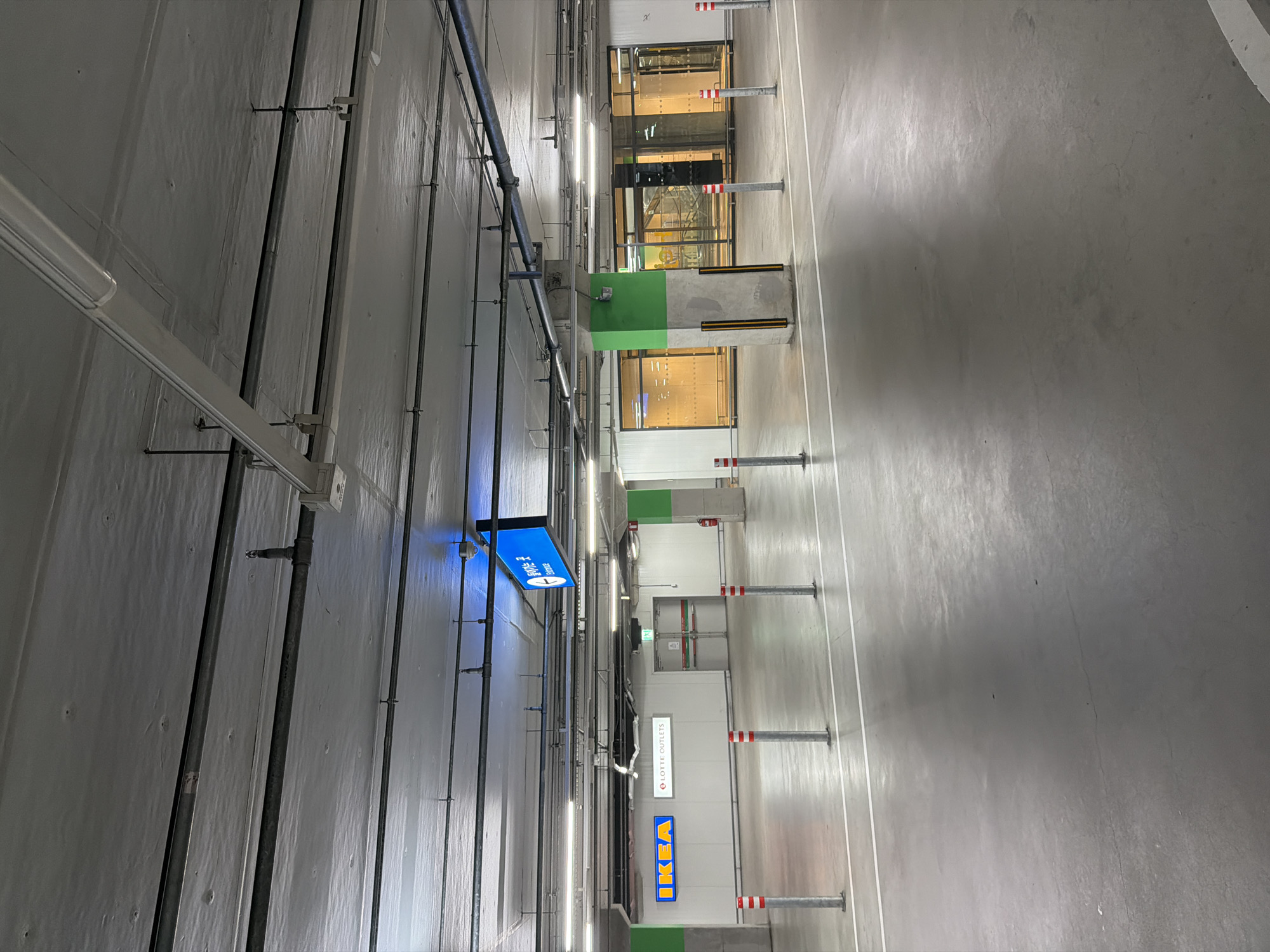 This is a photograph of a parking garage located at the Lotte Outlets and IKEA in Goyang-si, South Korea. The scene is illuminated by overhead fluorescent lights, casting a uniform glow on the concrete floor and ceiling. Several metal pipes and electrical conduits are visible running along the ceiling.
In the background, signs indicate "IKEA" and "LOTTE OUTLETS," with an "Entrance" sign above double doors. Pillars, some painted bright green, support the structure. Bolted to the floor at regular intervals are silver bollards topped with red and white reflective bands. The floor is a smooth, gray concrete surface with white painted lines marking parking bays or lanes. The overall impression is one of a functional, modern indoor public space. There are no people visible in the frame.