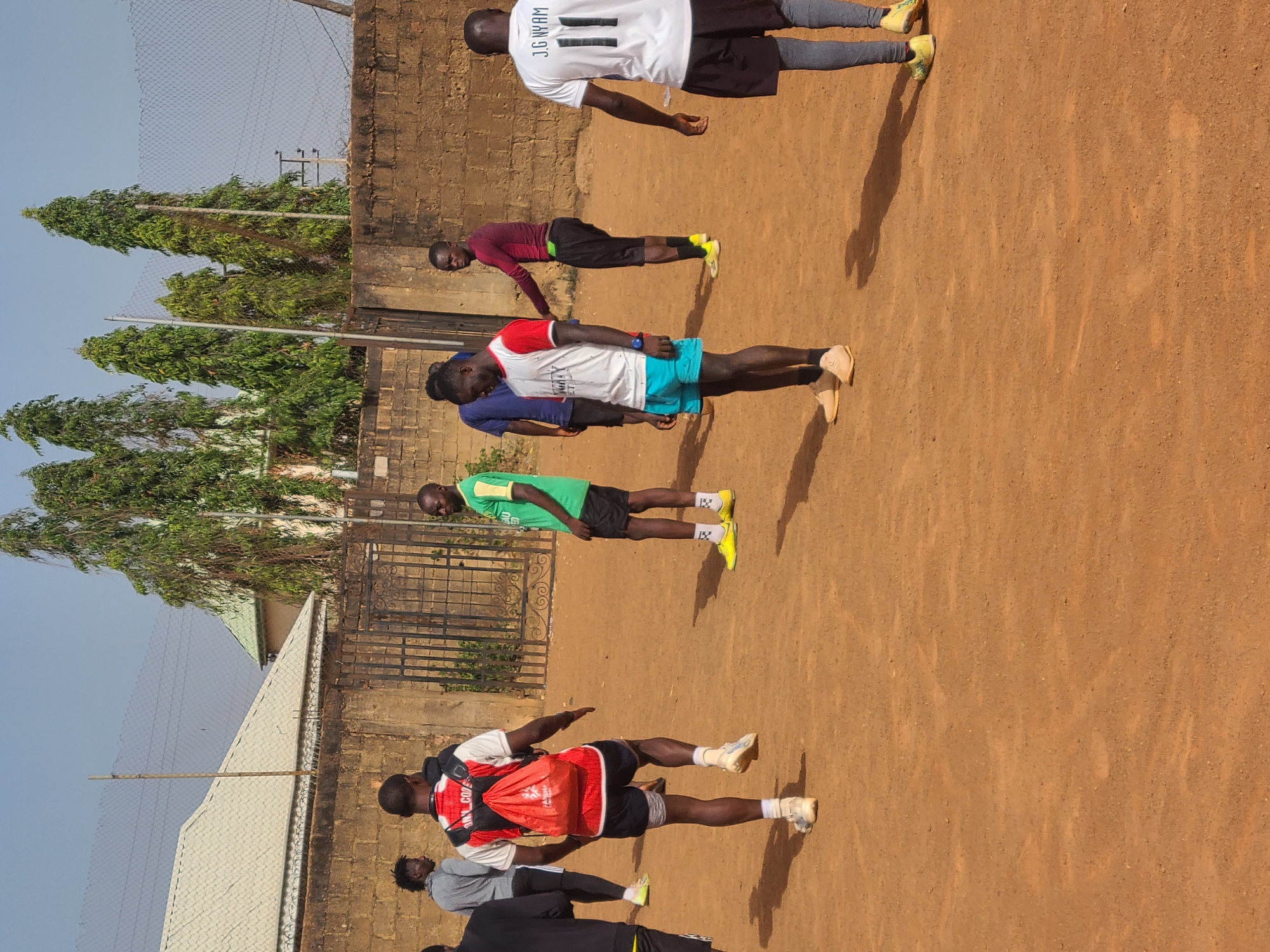 The image shows a group of seven men standing in a line on a dirt field in Barakin Lerawi, Nigeria. They appear to be stretching or warming up. The men are wearing athletic clothing including t-shirts, shorts, and running shoes. One man is wearing a red backpack. A brick wall runs along the side of the field with trees visible behind the wall. A fence is also visible along the top of the wall. The sky is clear and the lighting indicates it is daytime.