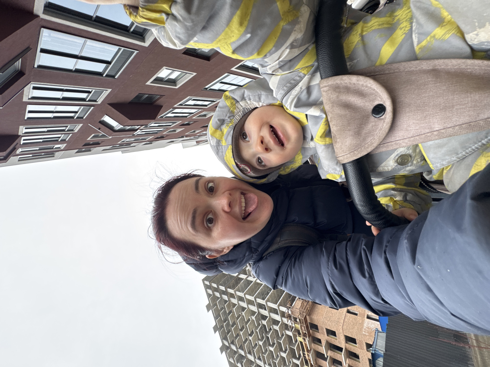 A rotated portrait-orientation photo captures a playful outdoor moment between an adult woman and a young child in Izhevsk, Russia. Both subjects are looking directly at the camera, suggesting a selfie.
The adult, positioned prominently in the lower left portion of the frame, has dark hair with reddish undertones and is wearing a dark blue puffer jacket, with a backpack strap partially visible. She is making a humorous expression, sticking her tongue out playfully.
To her right, a young child, likely a toddler, is visible, wearing a grey and yellow striped hooded winter suit or jacket over a light brown hat or hood lining. The child has a wide, happy grin and bright eyes, indicating joy and engagement with the camera. The child appears to be in a stroller or carrier, with a black handle and a beige padded safety strap visible in the foreground.
The background reveals an urban environment under an overcast sky, suggesting a cool or cold day. On the left, a modern, multi-story building with dark red-brown walls and numerous large, rectangular windows is visible. To the right, another building appears to be under construction, showing its raw concrete framework. The sky is uniformly grey and cloudy. The overall mood of the image is joyful and affectionate, focusing on the connection between the adult and child. There is no visible text in the image.