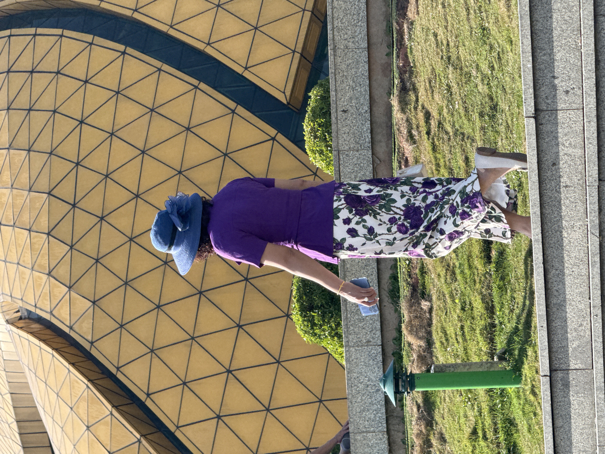 A person, wearing a purple top, a blue hat with a flower accent, and a floral skirt, walks on a paved walkway in front of a large, gold-colored, geodesic dome-like structure. The dome has a pattern of black lines forming triangles. To the left of the person, a green lamppost is visible. The ground is covered in green grass. The scene is located at the Truc Lam Zen Monastery in Da Lat, Vietnam. The person is holding a light blue smartphone in their right hand and is wearing white flat shoes.