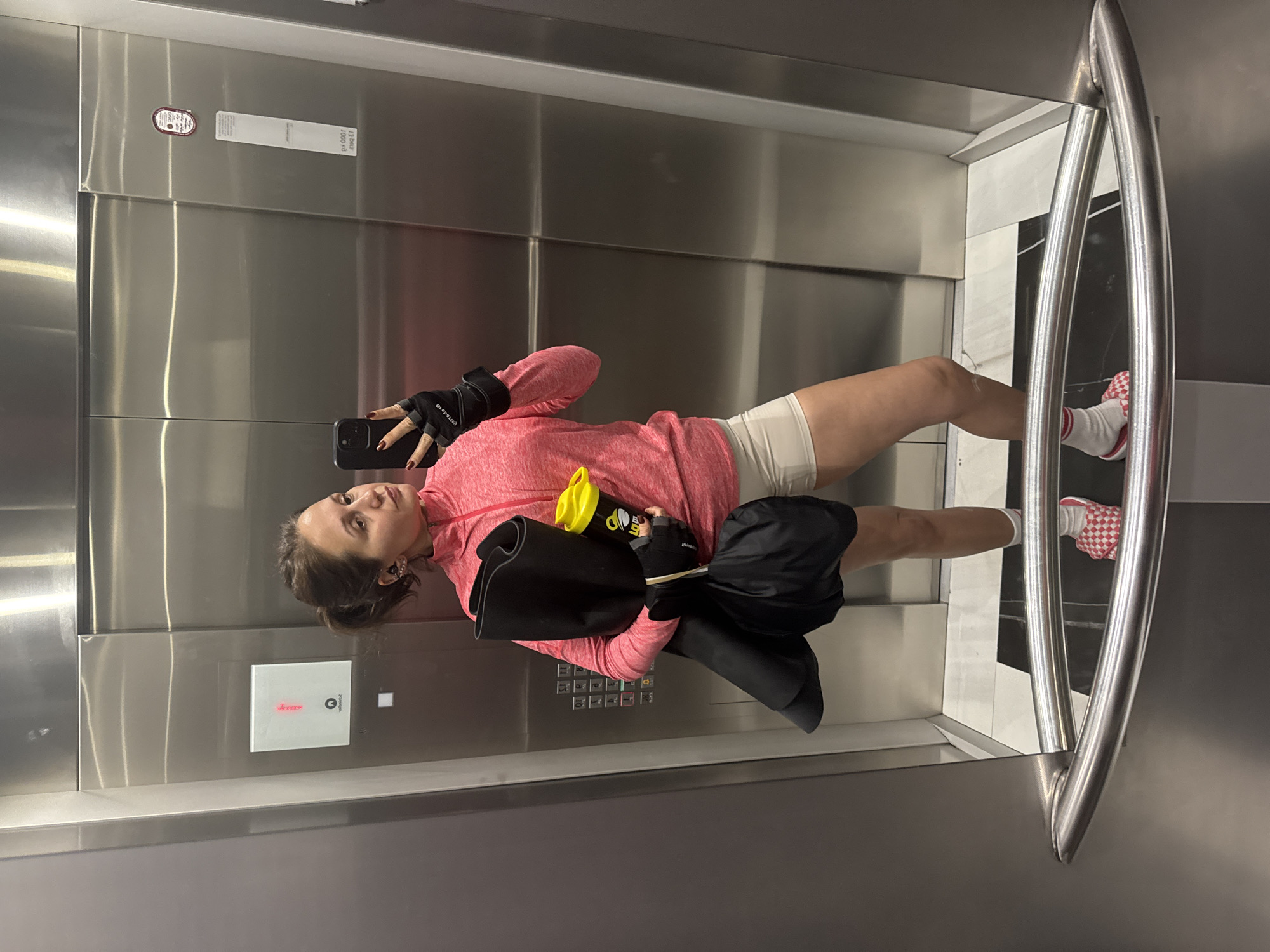 A woman is taking a selfie in an elevator. The elevator has stainless steel walls and a handrail. The woman is wearing a pink long-sleeved shirt, white shorts, gloves, and red and white checkered shoes. She is holding a water bottle and a black bag. The elevator is in Dubai, United Arab Emirates.