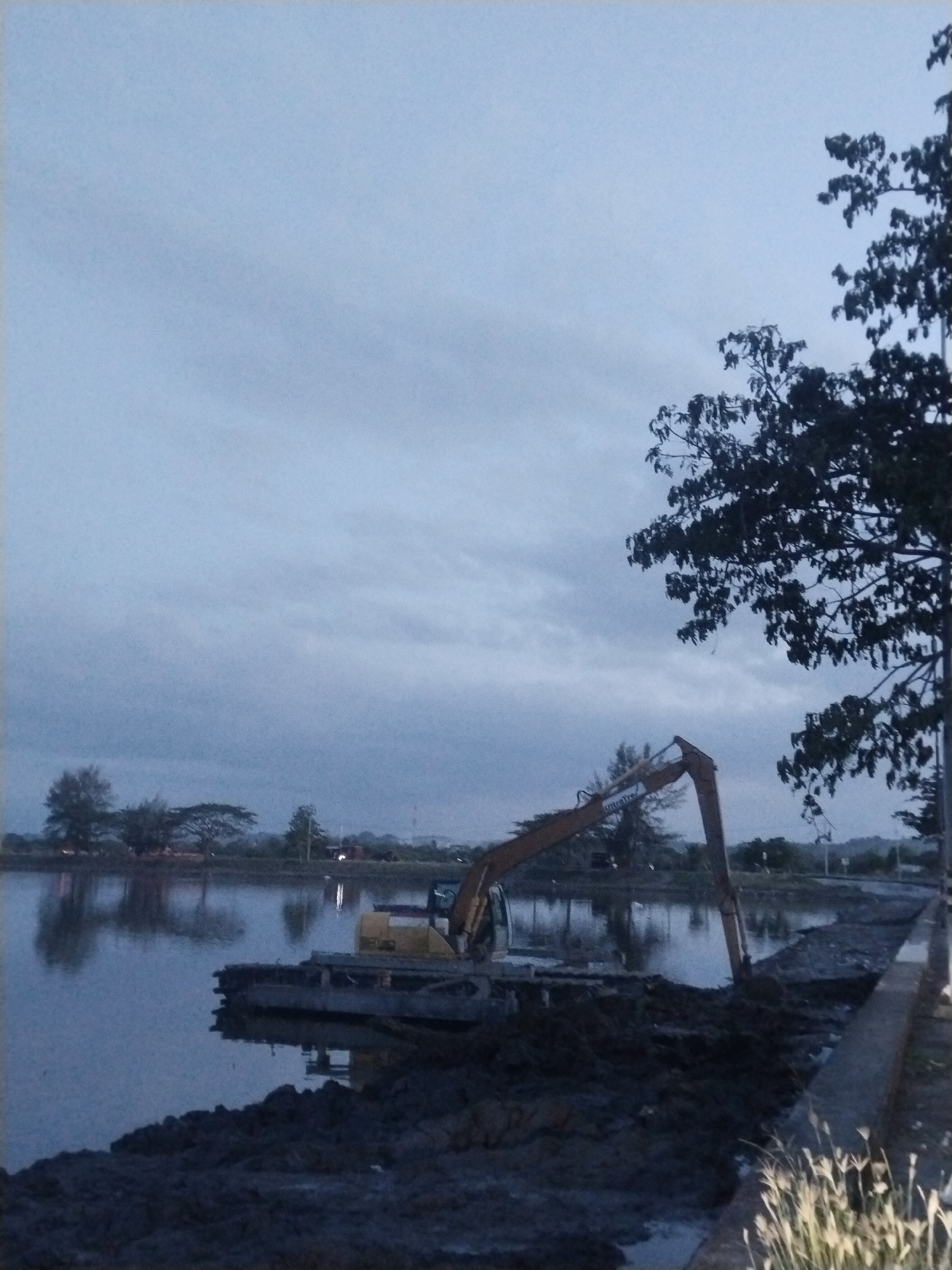 The image depicts a stationary excavator positioned on a barge, operating in a body of water adjacent to a concrete embankment in Lhokseumawe, Indonesia. The excavator arm is extended, suggesting dredging or excavation activities are in progress. Mud and sediment are piled up on the embankment. The water reflects the sky, indicating calm conditions. Silhouetted trees are visible on the opposite bank and a bare tree is in the foreground on the right side of the photo. The sky is overcast, suggesting either early morning or late afternoon light. Small white flowers grow near the embankment.