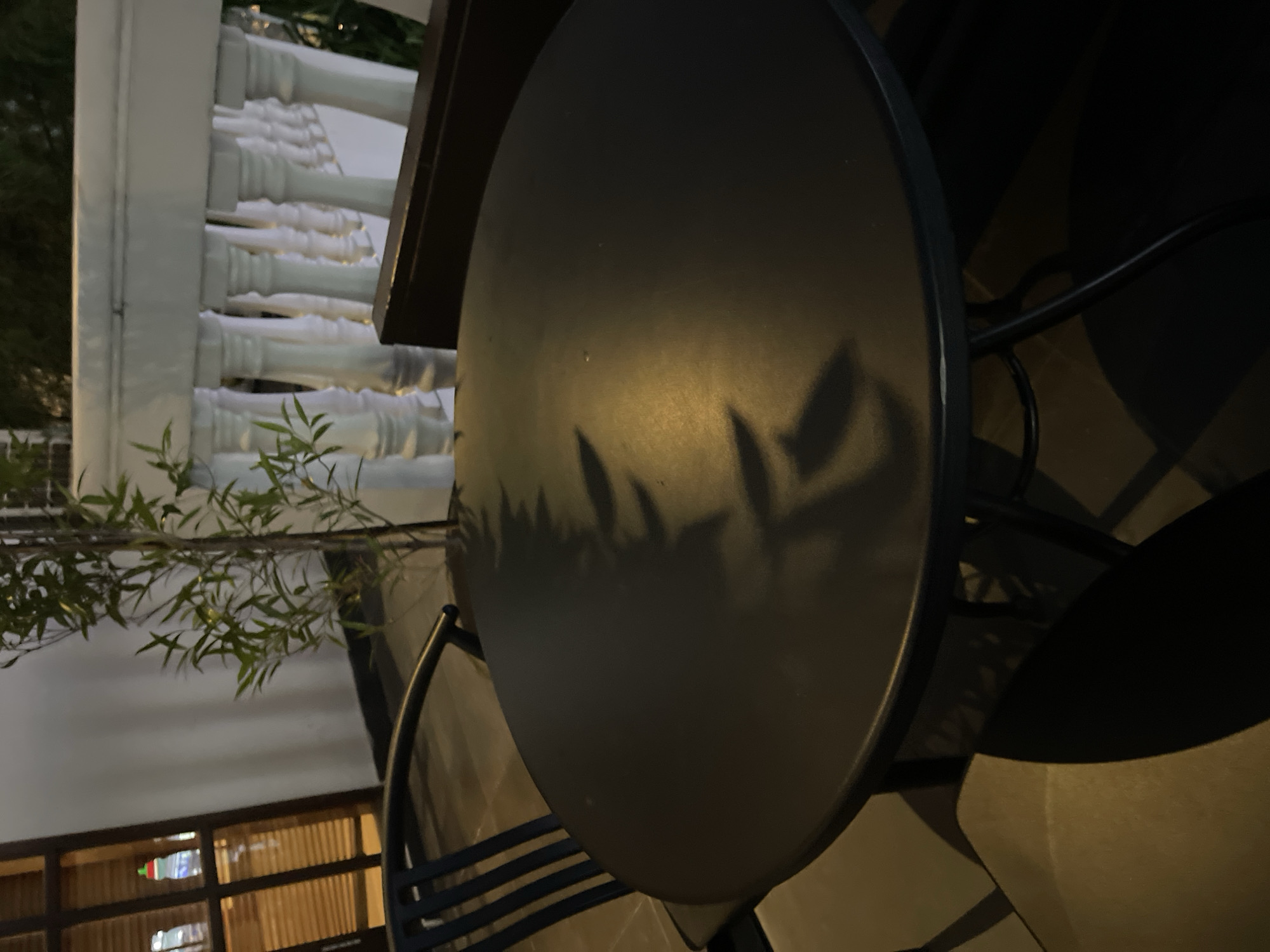 The image shows a round, dark-colored table with a shadow of foliage cast upon it, likely from an adjacent tree or bush. The table is part of a seating area, with chair backs visible around it. In the background, a white balustrade is partially visible, suggesting a terrace or balcony setting. Further back, glimpses of a building exterior can be seen with lit windows. The setting is likely a restaurant or outdoor seating area in North Kuta, Indonesia.