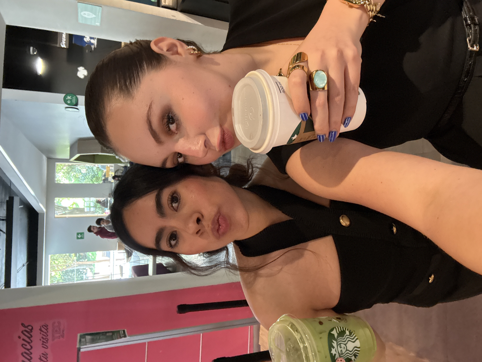 Two young adult females are depicted in a selfie taken indoors, specifically within a cafe or common area located on or adjacent to a Tecnológico de Monterrey campus in Mexico City, Mexico. The female positioned on the left has dark, wavy hair, wears a black strapless or off-the-shoulder top with gold buttons, and holds a clear plastic Starbucks cup containing a green beverage. She is looking directly at the camera with puckered lips. The female on the right has dark hair pulled back, wears a black top or blazer, and a dark belt. She holds a white Starbucks disposable coffee cup (400 ml) in her right hand. Her fingernails are painted with blue and white stripes. She wears multiple gold rings on her right hand, including a prominent one with a large turquoise-colored stone, and a gold bracelet. She is also looking directly at the camera with a slight pout. The immediate background on the left features a pink wall with partially visible text, including "Gracias" and "TEC." Large windows behind the subjects reveal outdoor trees and ambient daylight. In the blurred mid-ground, several other individuals are present within the establishment, including a male wearing a red hoodie. The setting features modern decor, including white and black wall sections, a dark ceiling, and a green directional arrow sign on a white wall.