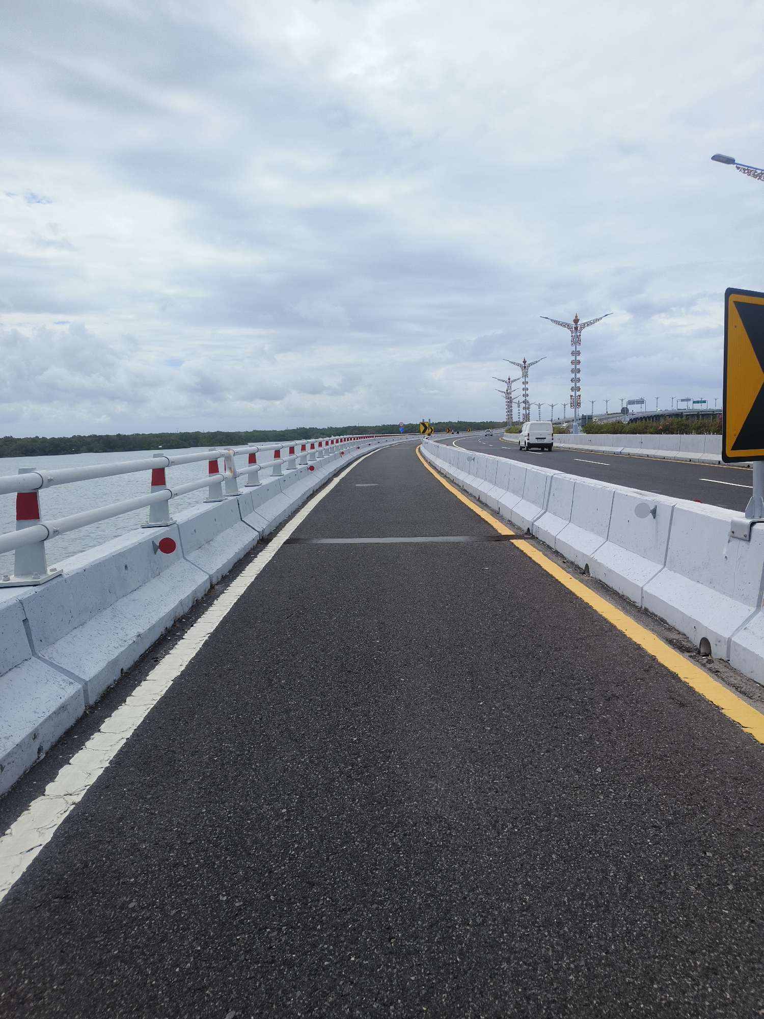 A long, empty asphalt road curves to the right, bordered by a white concrete barrier topped with a white railing and red and white posts. The railing is parallel to a body of water on the left, with a treeline visible in the distance. On the right side of the road, a yellow line demarcates the edge of the road from the concrete barrier, which is also seen further down the road. In the background, several tall, decorative streetlights with ornate tops are visible, along with a white van driving on the road. The sky is overcast with scattered clouds. The scene appears to be on a bridge or elevated roadway, potentially part of the Bali Mandara Toll Road in South Denpasar, Indonesia, which is known for its elevated sections over the sea and its distinctive decorative streetlights.
